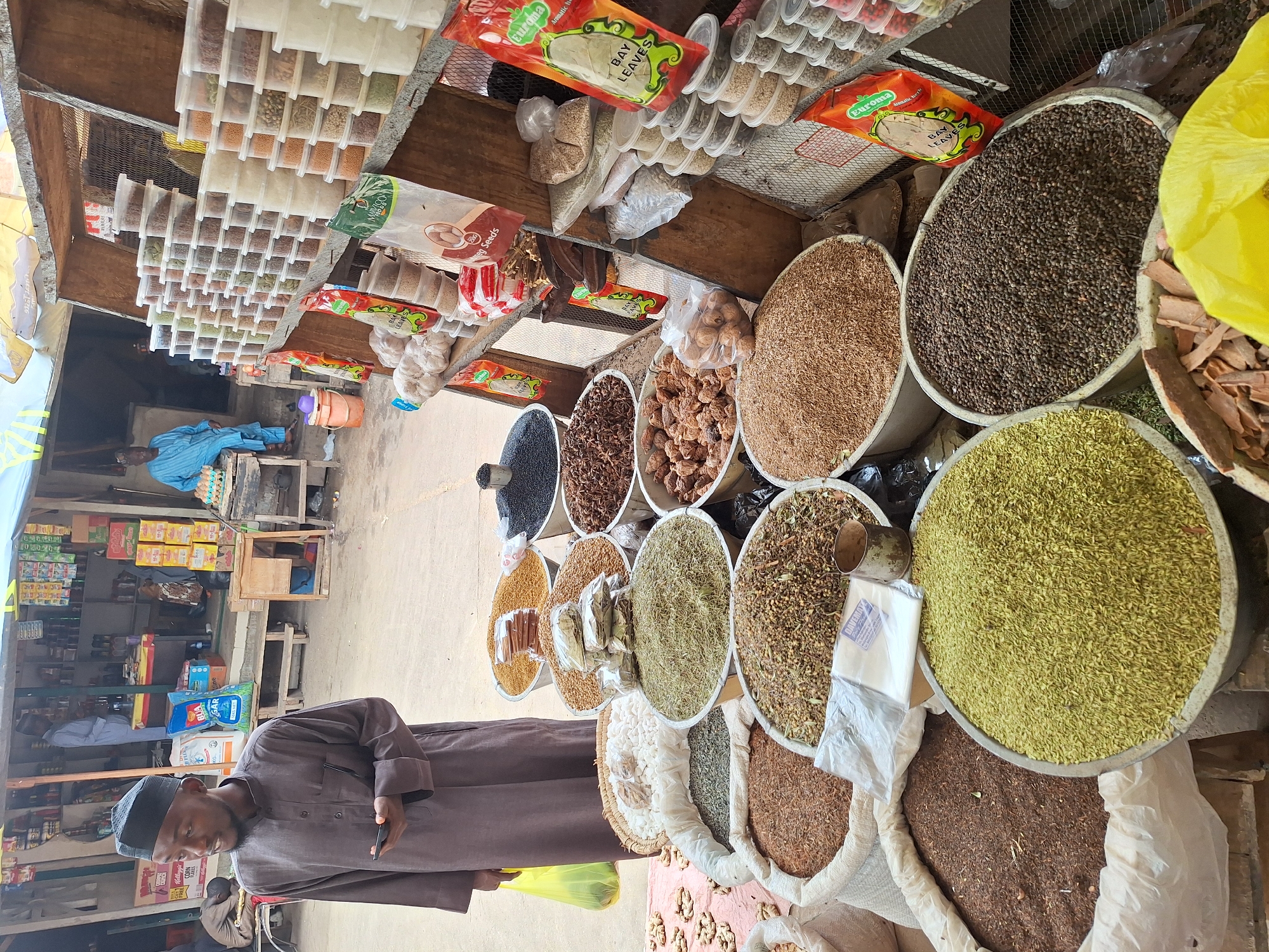 This image depicts a bustling market stall in Maiburiji, Nigeria, filled with a wide array of spices, herbs, and other goods. In the foreground, a man dressed in a dark brown traditional garment and a black cap stands, holding a smartphone. He is positioned in front of a colorful and abundant display of bulk ingredients.
The market stall features numerous large, circular bowls on the ground and on elevated surfaces, each filled with different dried products. Visible items include what appear to be black peppercorns, star anise, dried dates or similar fruits, various grains, green leafy herbs (possibly fennel or dried leaves), and other granular spices. Behind these bulk displays, a multi-tiered rack holds an assortment of spices and seeds in clear, small rectangular containers.
Packaged goods are also prominent, with several red and green "Euroma BAY LEAVES" pouches visible, alongside a package labeled "Reg Seeds." Further into the stall, a large white bag with blue text, possibly reading "BUA SUGAR," can be seen. A bright yellow plastic bag hangs near the foreground man.
In the background, another man in a light blue outfit is seated near a tray of eggs, while a third person in a light grey shirt is partially visible further back. The scene is brightly lit, indicating daytime, and the overall atmosphere suggests a lively commercial setting with rustic market structures.