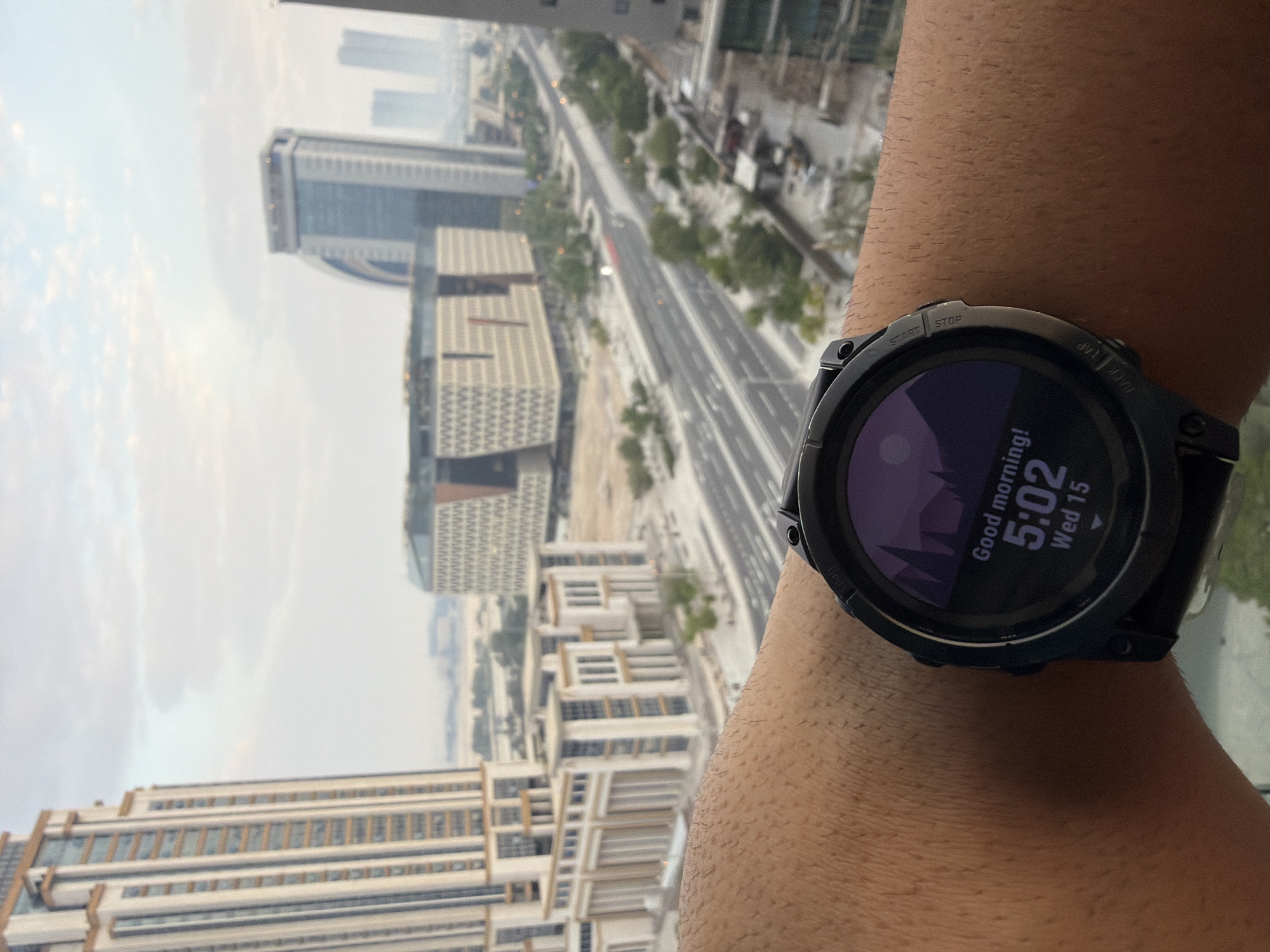 The image shows a close-up of a person's forearm wearing a smartwatch, with a cityscape in the background. The smartwatch displays "Good morning! 5:02 Wed 15" on its screen, with a minimalist mountain graphic. The background reveals a modern city with several tall buildings, including a distinctive curved building, and a multi-lane road. The sky is overcast. The perspective suggests the photo was taken from a high vantage point, likely a building in Lusail, Qatar, given the architectural style of the buildings which are characteristic of the city.