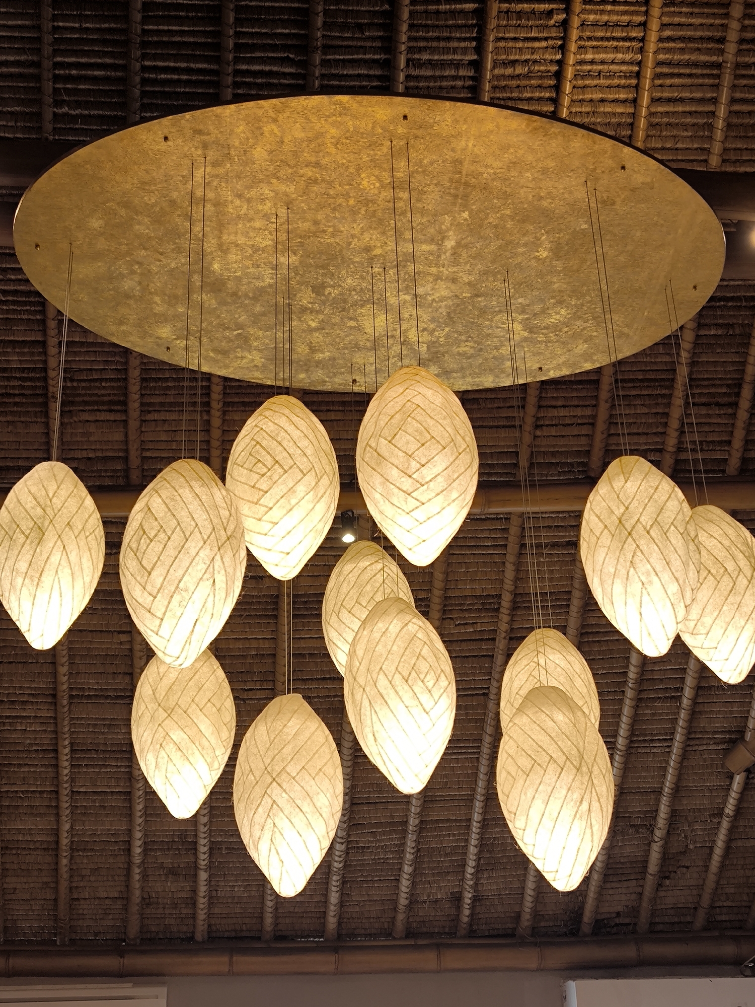 The image shows a lighting fixture hanging from a ceiling. The ceiling is covered in dark, thatched material. Above the hanging lights is a large, circular, golden-colored disc. Suspended from the disc are multiple oval-shaped lanterns. The lanterns are made of a textured, light-colored material and are illuminated. The photo appears to have been taken indoors in Mekarsari, Indonesia.