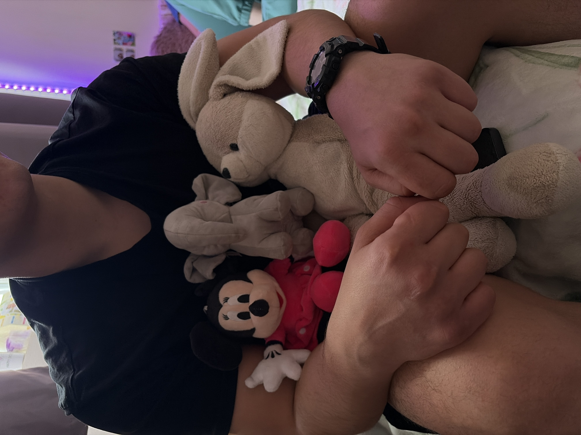 A person is sitting down, holding a beige plush rabbit, a grey plush elephant, and a red plush Mickey Mouse doll. The person is wearing a black t-shirt and a black digital watch. The setting is indoors, with a bed in the background with a patterned duvet and ambient light. No information suggests the location is Sofia, Bulgaria.