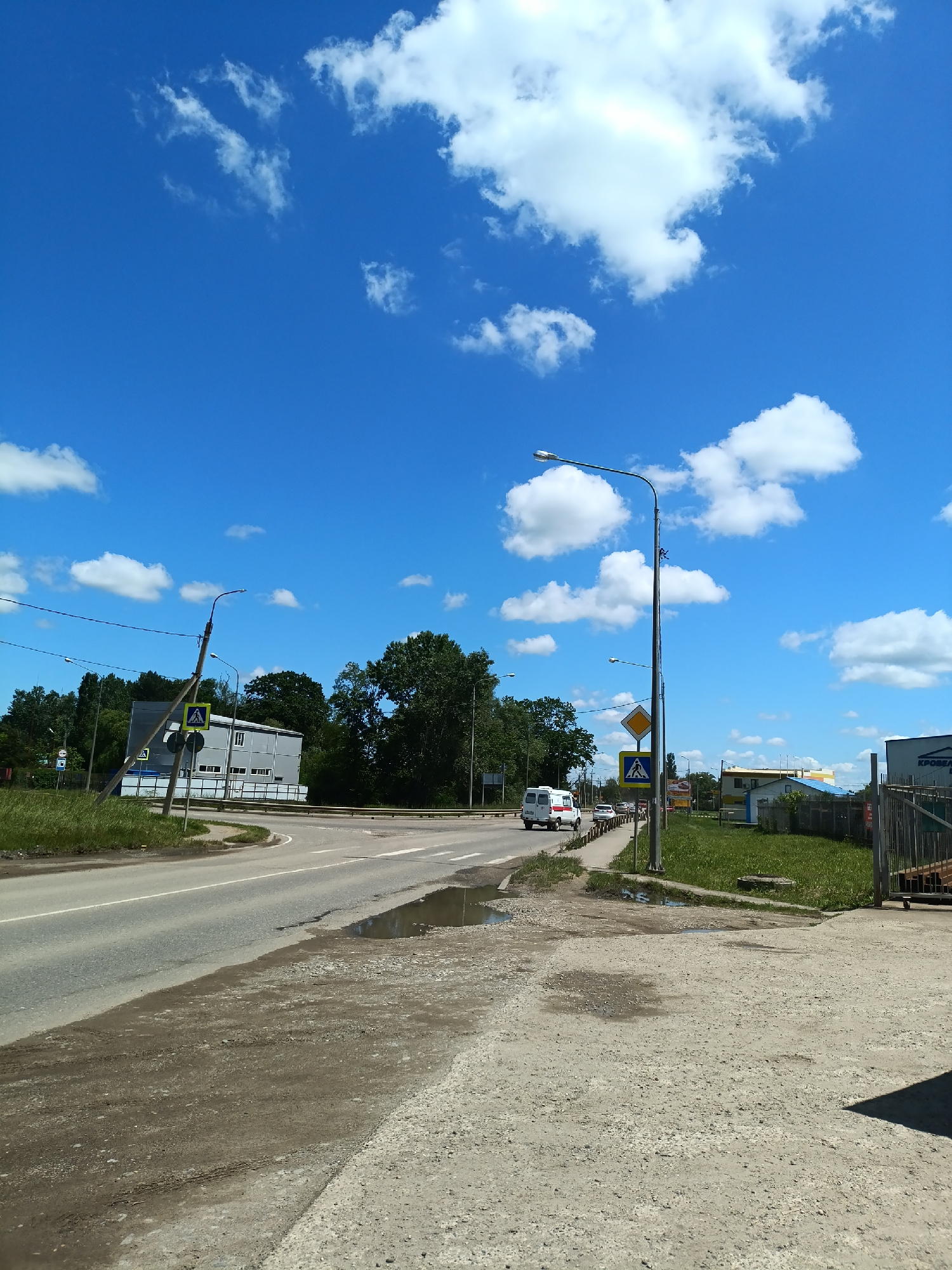 This image captures a bright, sunny day scene at a road intersection or junction in Goryachiy Klyuch, Russia. The sky is a vivid blue, adorned with scattered, fluffy white cumulus clouds.
In the foreground, a rough dirt and gravel shoulder, marked with tire tracks and holding several small puddles suggesting recent rain, transitions into an asphalt road. The road features white line markings and a clearly visible pedestrian crossing marked by a yellow triangular sign with a walking person. Several streetlights stand tall, along with a yellow diamond-shaped sign, typically indicating a main or priority road.
To the left, the road curves past a modern, grey industrial-style building with multiple rectangular windows. Lush green trees and grass flank the roadside, creating a natural backdrop against the man-made structures. A metal guardrail runs alongside a section of the road.
On the road, a white van, possibly an ambulance or medical transport due to visible red crosses on its side, is seen moving away from the viewer. Other vehicles are discernible further down the road. To the right, another pedestrian crossing sign is visible, and beyond it, a grassy area hosts a small blue shed-like structure and a more distant building. A partially visible sign near the right edge of the frame reads "КРОВЕЛЬНЫЕ" (Roofing), indicating the presence of commercial businesses. The immediate ground on the right is a concrete or gravel surface, with a shadow extending into the frame.
The scene depicts a typical semi-urban or industrial area, vibrant with daylight, and conveys a neutral, functional atmosphere. No specific human activity or prominent emotions are visible.