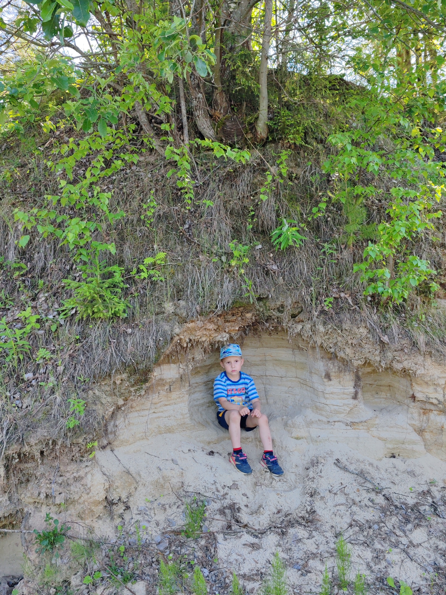 A young boy is sitting in a natural alcove formed by layered sandy soil, likely a cliff face or embankment, in the city of Chukhloma, Russia. The boy is wearing a striped blue and white t-shirt, dark shorts, a blue cap with a pattern, and athletic shoes with pink accents. He is positioned towards the right side of the frame, with his legs extended. The alcove he is sitting in reveals distinct horizontal layers of sediment. Above and behind the alcove, the embankment is covered with dense green foliage, branches, and dry grasses. The ground in front of the alcove is composed of sand and scattered debris. The lighting suggests it is daytime, with sunlight filtering through the trees.