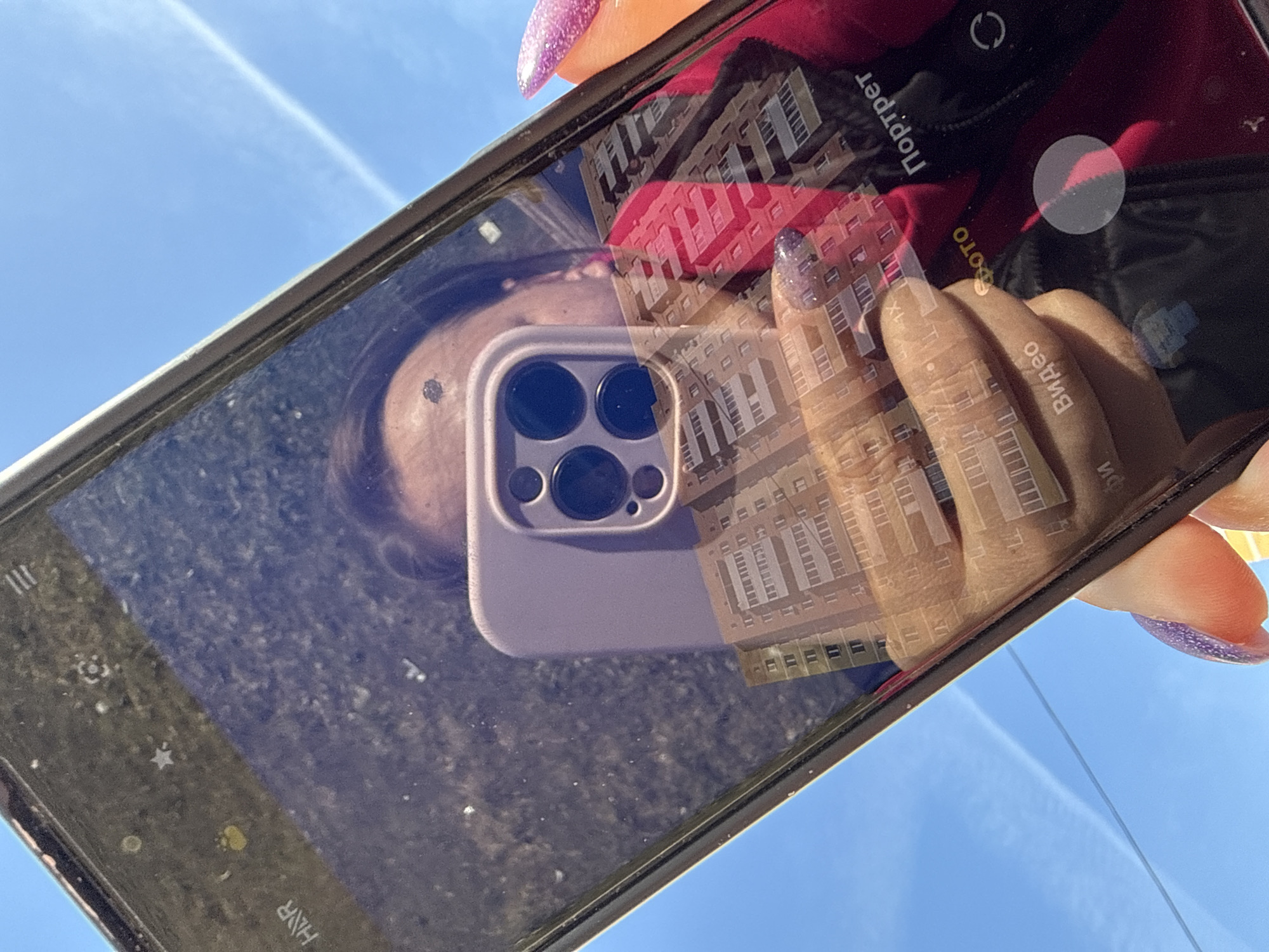 An individual is holding a smartphone. The smartphone's screen displays a reflection of a building. The reflection and the surrounding environment show clear, sunny weather. The person's hand with decorated nails is visible holding the phone. The phone is pointed towards the sky, with what appears to be a contrail of an airplane. Based on the building reflection, the location is in Dmitrov, Russia.