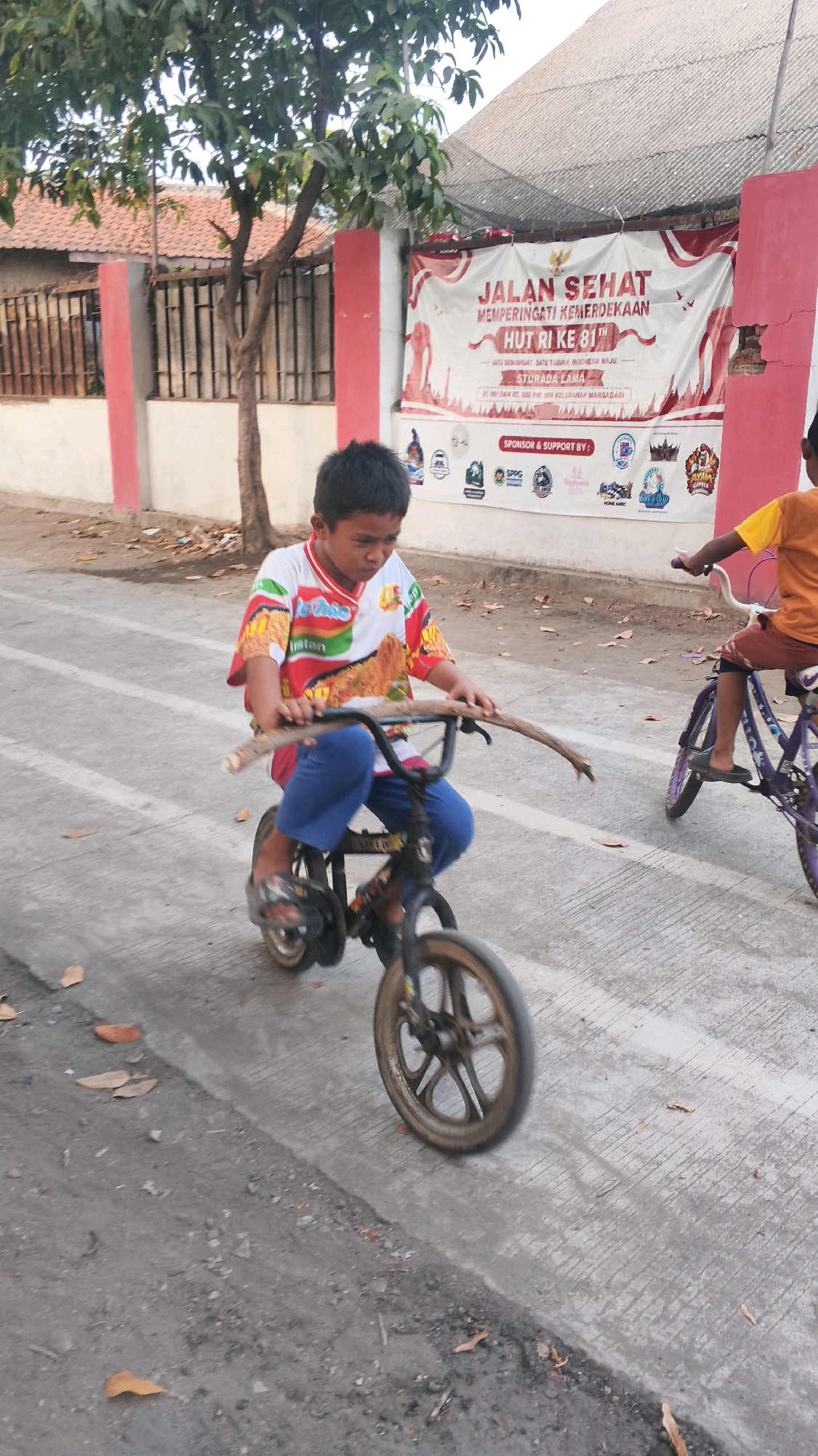 In Indramayu, Indonesia, a young boy rides a bicycle down a paved street. He wears a colorful t-shirt and blue pants, gripping a long stick as if it were handlebars. The bicycle is small and dark-colored with a single wheel visible. In the background, a red and white banner hangs across a building, reading "JALAN SEHAT MEMPERINGATI KEMERDEKAAN HUT RI KE 81" which translates to "Healthy Walk Commemorating the 81st Independence Day of the Republic of Indonesia." Another child on a purple bicycle is visible to the right. The street is lined with trees and buildings, suggesting a residential or community area. The ground is paved with concrete and scattered with fallen leaves.