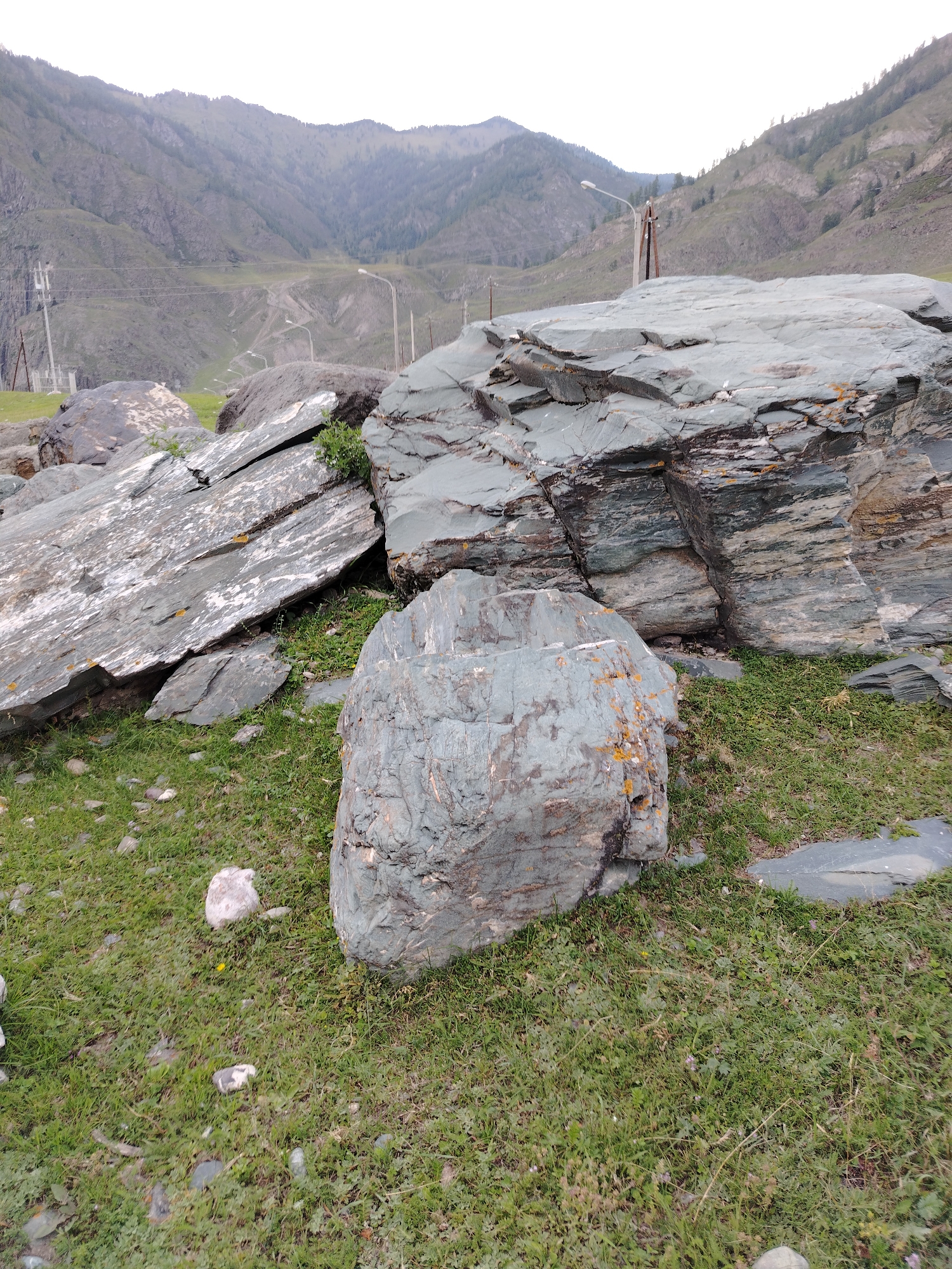 A natural scene is depicted, featuring large, grey, weathered rocks scattered across a grassy terrain. The rocks exhibit layered textures and some are tinged with orange and yellow mineral deposits. In the background, a mountainous landscape with green slopes is visible, partially obscured by a hazy sky. A road with streetlights winds through the valley in the distance. Several power poles and electrical wires are also present, suggesting proximity to human infrastructure. The overall impression is one of a rugged, untamed natural environment.