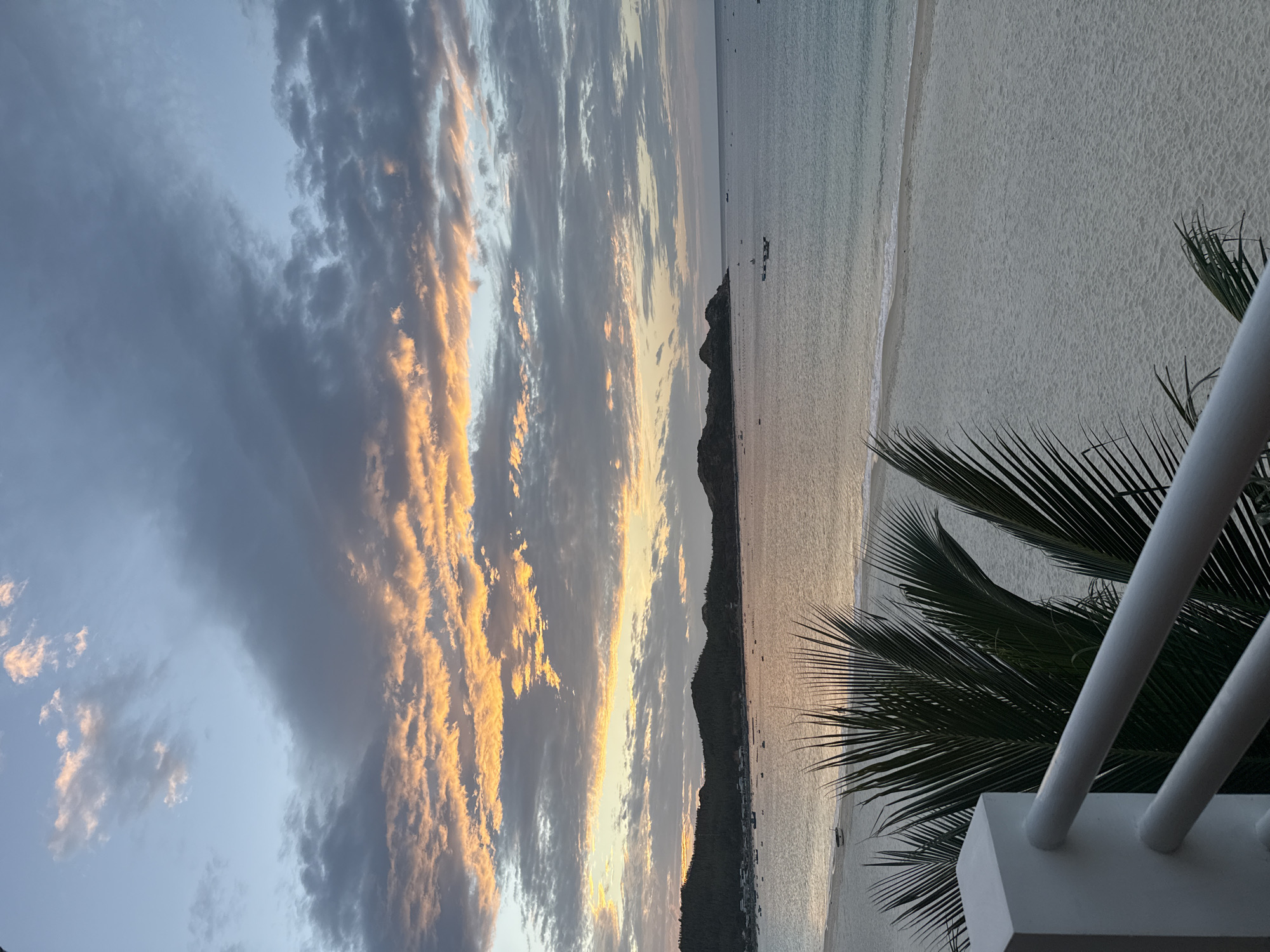 This image captures a serene coastal sunset scene in Song Cau, Vietnam. The foreground features a white railing and lush green palm fronds, partially obscuring a pristine white sandy beach. Beyond the beach, the calm ocean stretches to a horizon dotted with small boats. The background is dominated by a range of green, undulating hills. The sky above is a canvas of dramatic sunset colors, with streaks of orange and yellow clouds contrasting against shades of gray and soft blue. The overall impression is one of peaceful natural beauty.