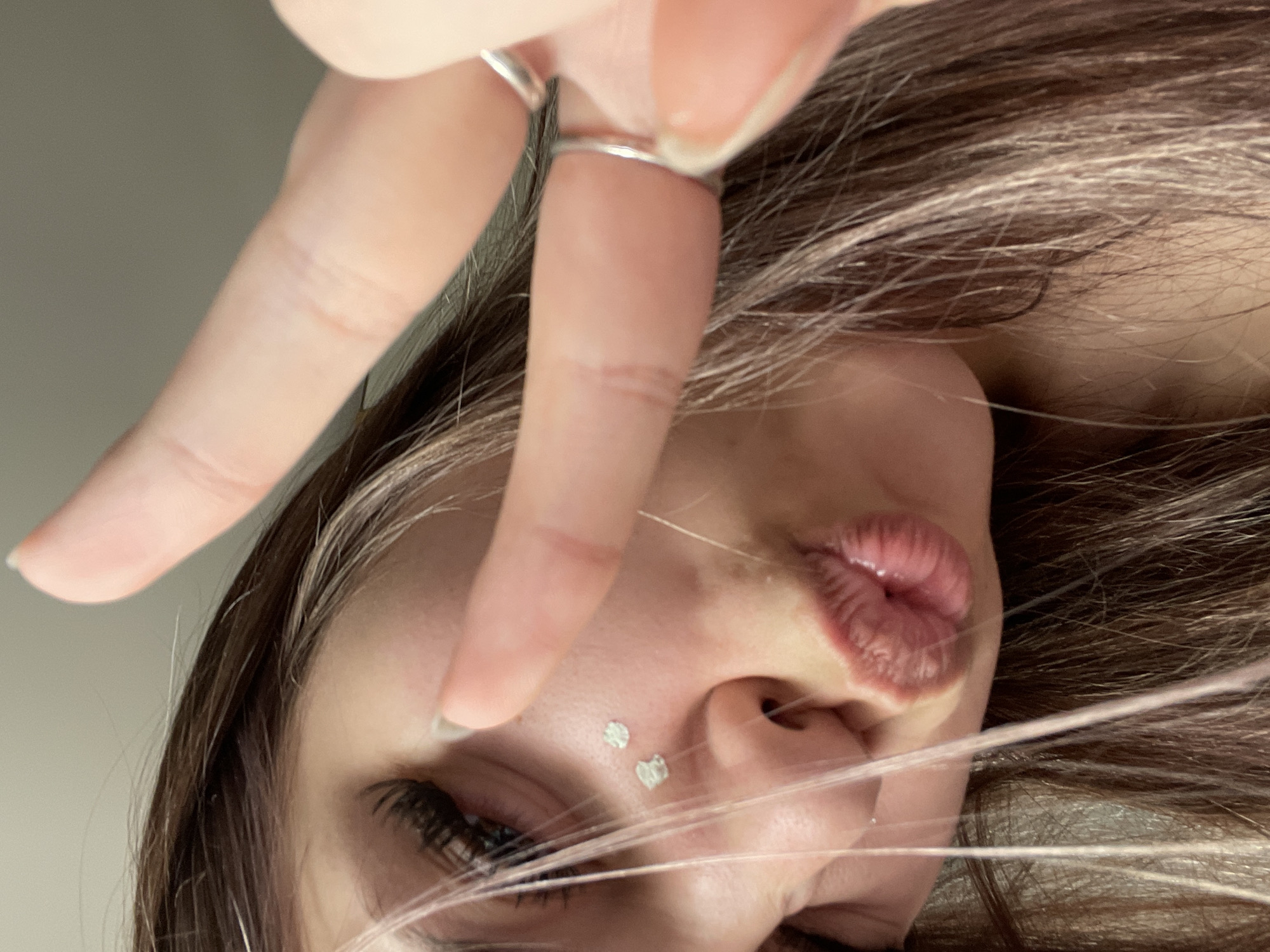 A close-up, tilted selfie captures a young individual, likely female, from a low angle. The subject's right eye with prominent lashes, nose, and full, slightly pursed lips are visible. Two small, shiny, heart-shaped decorations are affixed to her upper cheek/side of her nose. Her long, dark brown hair, streaked with lighter highlights, frames her face, with several strands extending across the foreground.
A hand, adorned with two silver-colored rings, is positioned in the upper left, making a peace sign gesture. The background is a plain, light-colored, out-of-focus surface, suggesting an indoor setting. The image conveys a casual and playful mood. There is no visible text. The location context provided is Saint Petersburg, Russia.