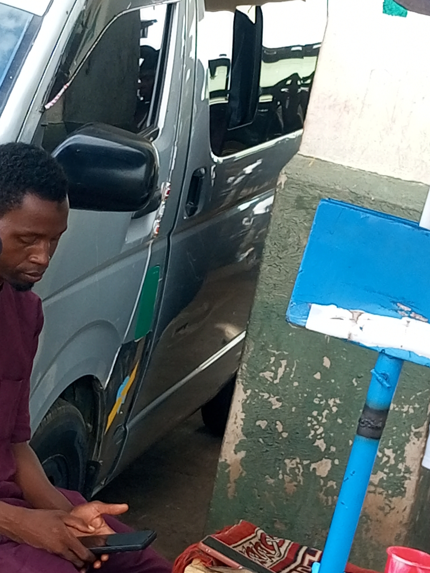 A Black man is sitting next to a grey minibus and holding a mobile phone. He is wearing a burgundy tunic. He is looking down at the phone. The minibus has a black side mirror. A blue pole is visible to the right of the man, with a blue rectangular object attached to its top. The background consists of a green, textured wall with white flecks. The scene appears to be in an outdoor setting in Gombe, Nigeria.