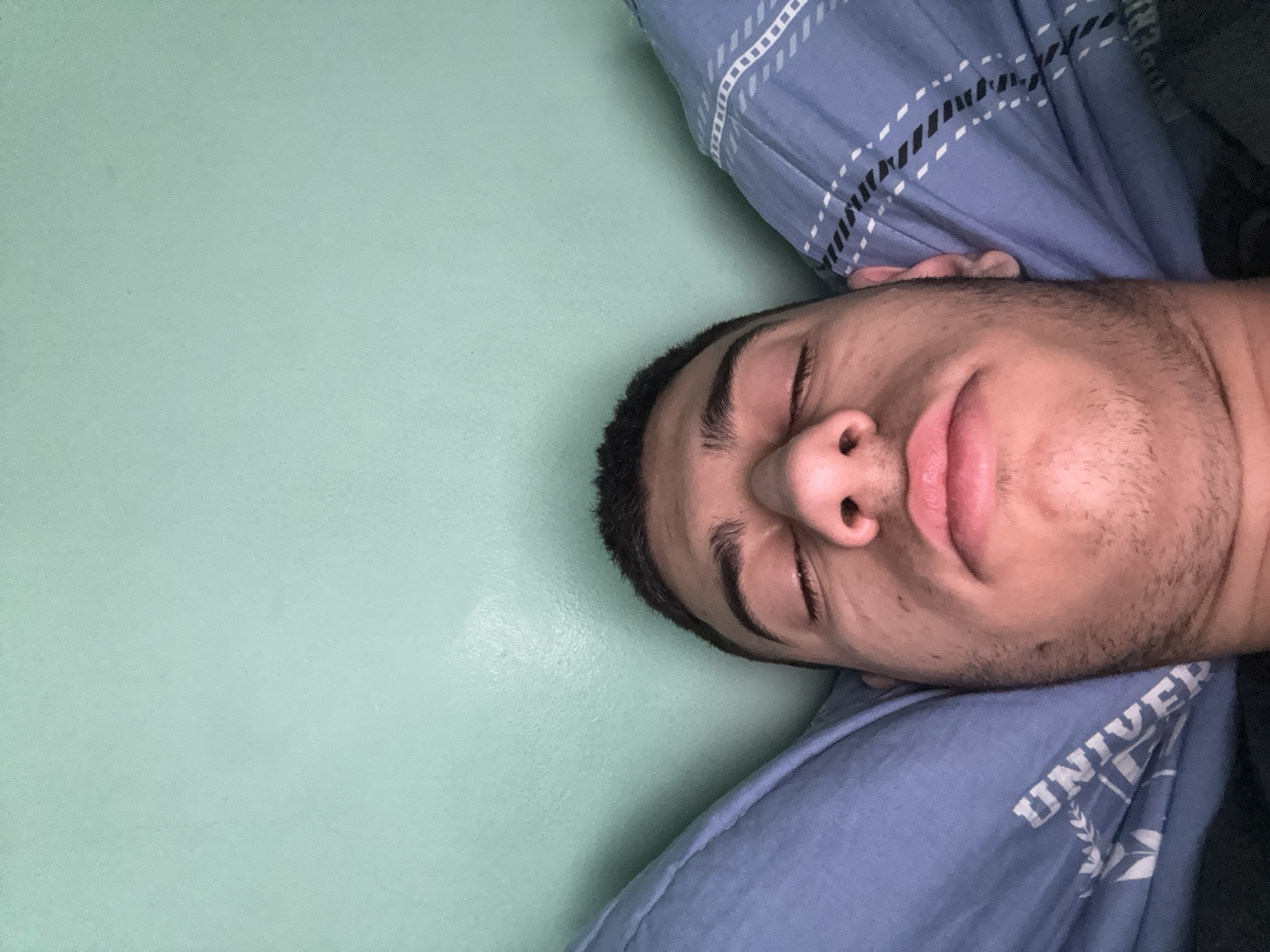 A close-up, slightly angled shot shows a man lying on his back. He has dark hair, closed eyes, and is resting on a blue pillow with a white and black checkered pattern. He is wearing a blue shirt with the word "UNIVER" visible. The background is a light green, possibly a wall. The location cannot be determined to be Sofia, Bulgaria, without additional information.