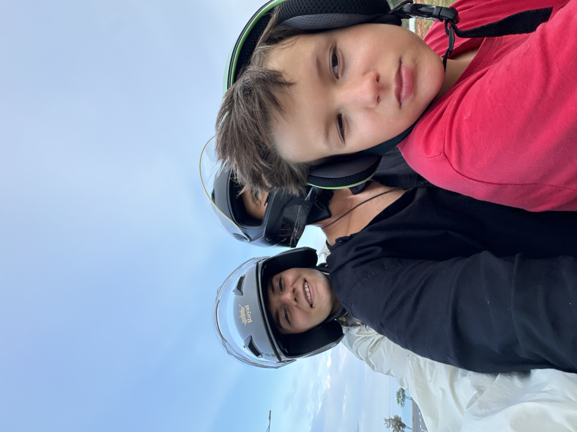 A selfie-style close-up photograph, rotated counter-clockwise, features two individuals wearing helmets against a bright, slightly cloudy blue sky. In the foreground, a child or young person with brown hair wears a green and black open-face helmet, equipped with what appears to be an integrated communication headset. They are wearing a red short-sleeved shirt and look directly at the camera with a neutral, slightly pensive expression.
Behind the child, and partially obscured, is an adult, also wearing an open-face black helmet with a clear visor. This helmet has the word "Royal" and a sun logo visible on its side. The adult is smiling warmly at the camera and wears a black top, possibly with a light-colored jacket or clothing underneath. Wires from a communication device are also visible near the adult's helmet.
The background shows a vast expanse of light blue sky with faint clouds, suggesting daytime. In the lower right corner, glimpses of what might be a road and distant trees or coastal vegetation are visible, indicating an outdoor setting. The overall impression is of two people on a motorcycle or scooter, possibly taking a break or in transit. The location is Mũi Né, Vietnam.