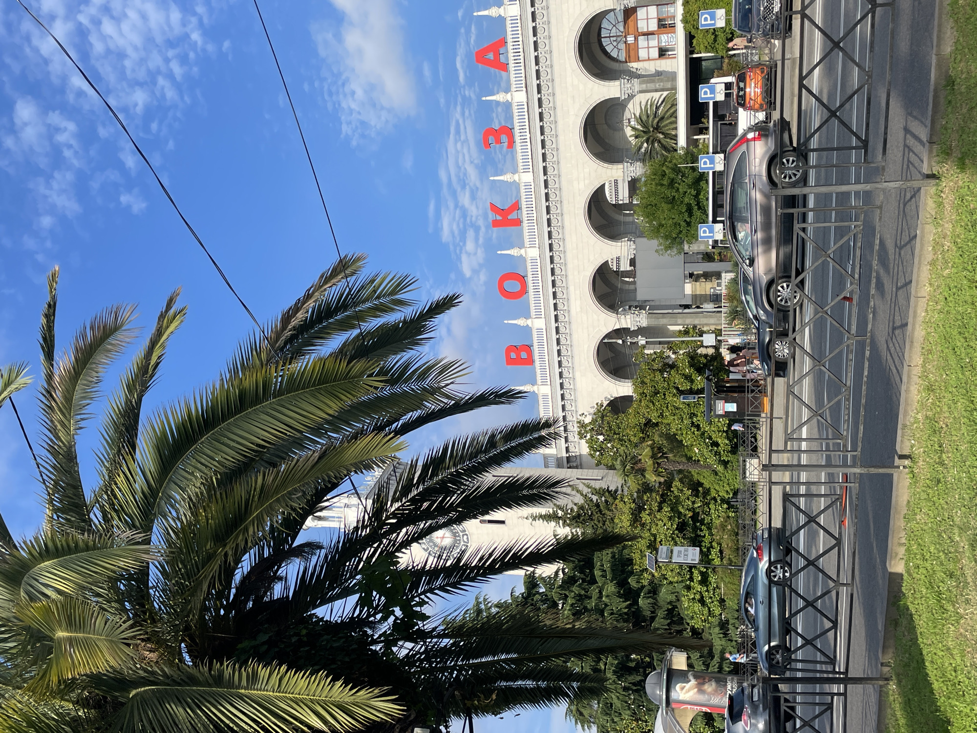 The image shows a street scene in Sochi, Russia, with a prominent railway station building in the background. A large palm tree with green fronds partially obscures the view. The railway station features a clock tower and is adorned with red lettering that spells out "ВОКЗАЛ" (VOKZAL), which means "station" in Russian. The building has a classical architectural style with arched entrances and decorative elements. In the foreground, cars are visible on the road, with a metal barrier separating the sidewalk from the street. The sky is blue with scattered white clouds. The overall atmosphere suggests a sunny day in a city with a subtropical climate.
