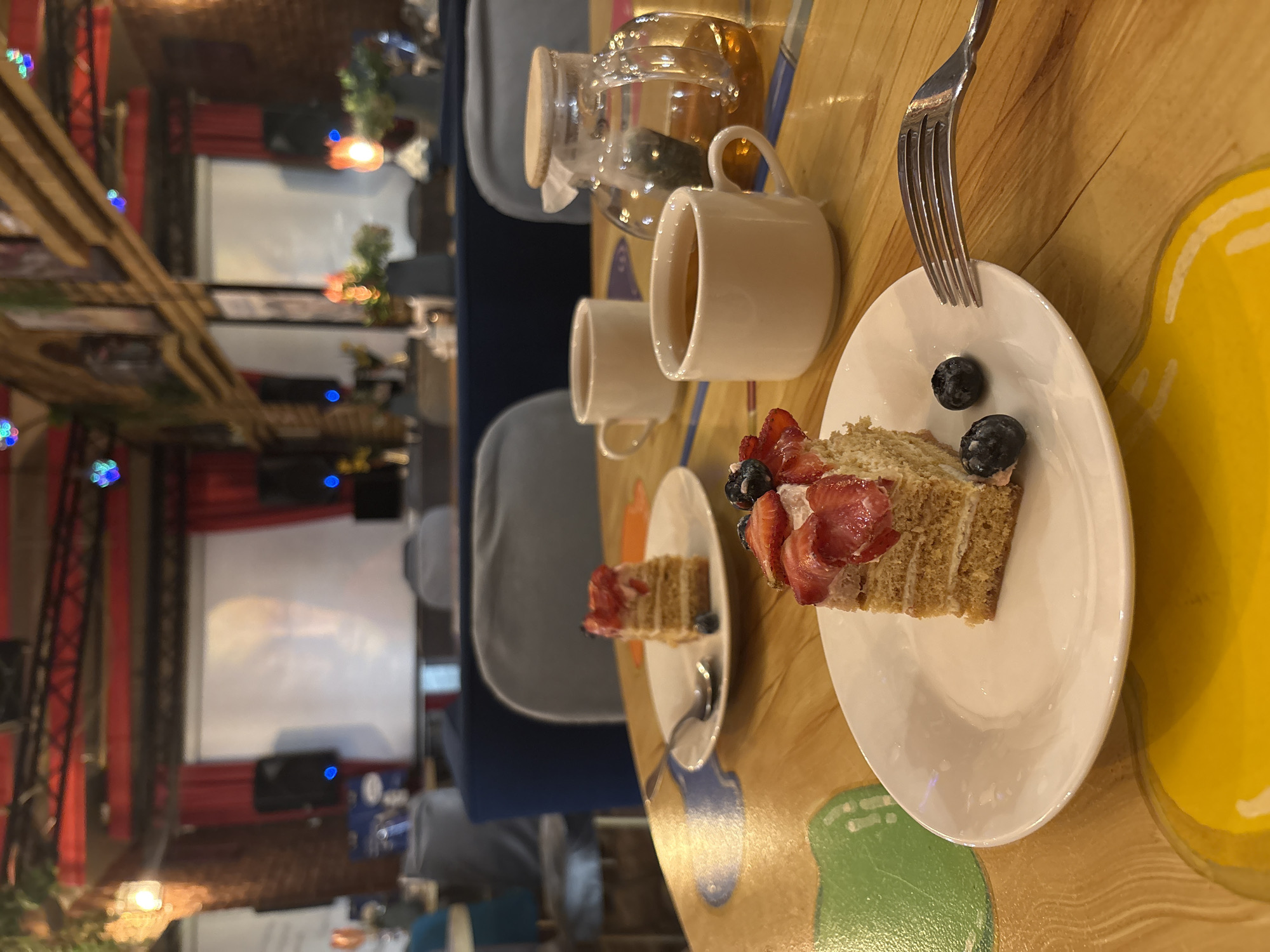 The image captures a close-up view of two slices of layered cake topped with strawberries and blueberries, served on white plates. One slice is in the foreground on the right, accompanied by a fork. In the middle ground, another slice of cake sits on a plate with a spoon. Two white teacups and a glass teapot filled with amber liquid are also present on a wooden table with colorful circular designs. The background reveals a dimly lit interior with plush seating, a projector screen displaying a blurred image, and industrial-style red beams and lighting fixtures, suggestive of a cafe or bar in Saint Petersburg, Russia. The overall atmosphere is cozy and inviting.