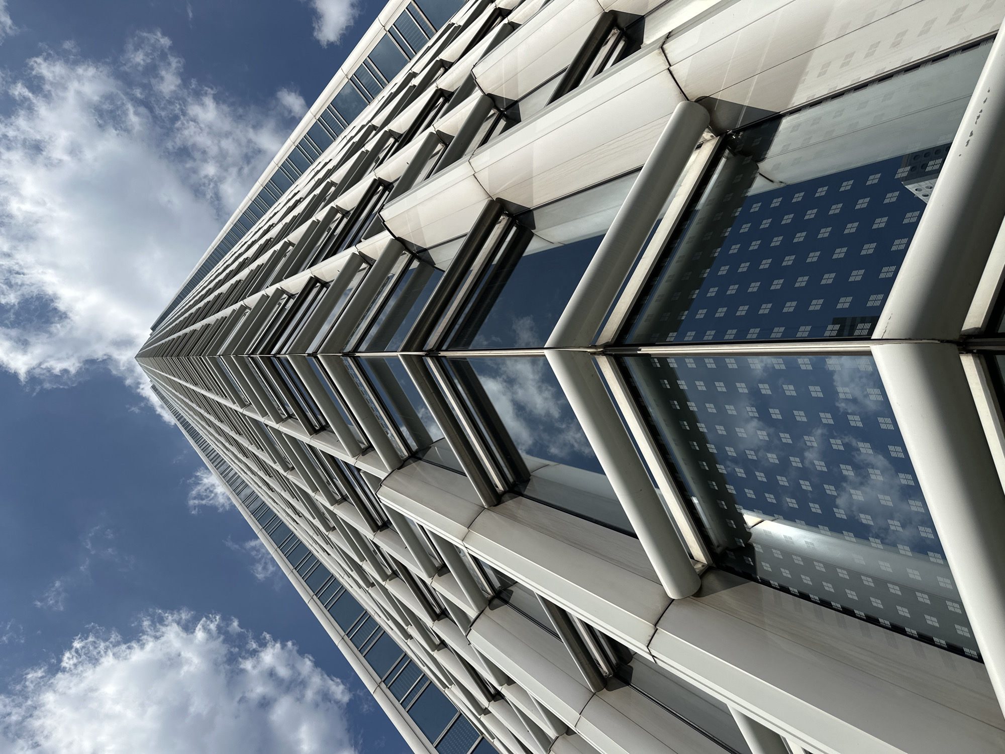 This is a low-angle, worm's-eye view photograph of a modern skyscraper in Hong Kong, China. The building features a white, angular facade with a grid of windows. The sky above is blue with scattered white clouds. The glass windows reflect the sky and other buildings, including what appears to be another skyscraper with a grid pattern of windows. The building's structure is composed of interlocking geometric shapes, creating a sharp, upward-pointing perspective. The image captures the scale and architectural style of a contemporary urban environment.