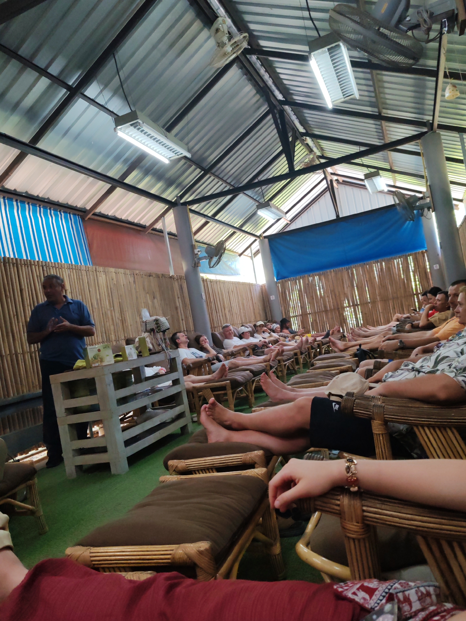 This image captures a foot massage or spa session taking place in an open-air or semi-covered pavilion in Ban Khlong Khian, Thailand. A group of approximately ten people, mainly adults of various ages, are seated in a row of natural bamboo/rattan reclining chairs with brown padded cushions, their legs extended onto footrests. They appear relaxed and comfortable, some observing their surroundings, others possibly conversing.
On the left, a local man in a dark blue polo shirt stands beside a light-colored wooden multi-shelf table, gesturing with his hands as if explaining or instructing. The table holds several small bottles and other items, though specific text on them is unreadable due to blurriness.
The setting features walls made of vertical bamboo poles, lending a rustic, tropical aesthetic. The roof is constructed from corrugated metal sheets supported by dark metal beams. Several fluorescent strip lights illuminate the space, complemented by multiple wall-mounted oscillating fans and some ceiling-mounted fans, suggesting a warm climate. A blue and white striped awning or curtain is visible on the left wall, with a plain blue tarp covering another section in the background. The floor is covered with green turf or carpet.
The perspective is from one of the chairs, looking across the room towards the other participants and the standing attendant. Notable details include a "NIKE" logo visible on a person's black shorts and a decorative, multi-stone bracelet on a person's wrist in the foreground. The overall atmosphere conveys a tranquil and communal spa experience.