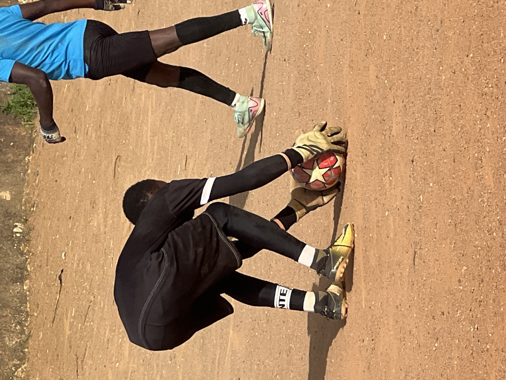 Two young males are in a dusty, outdoor area, likely a field. The male in the foreground is a goalkeeper, crouched low, wearing a black jersey and shorts, black full-length leggings, white and black socks with "NTE" printed on them, and yellow cleats. He is reaching for a red and white soccer ball with his gloved hands. The second male, to the right and slightly behind, is wearing a blue t-shirt, black shorts, black knee-high socks, and light green and pink athletic shoes. He appears to be walking. The ground is a sandy, reddish-brown dirt with small pebbles and some scattered vegetation in the background. The scene is consistent with a soccer practice or informal game on a dirt field in Jos, Nigeria.