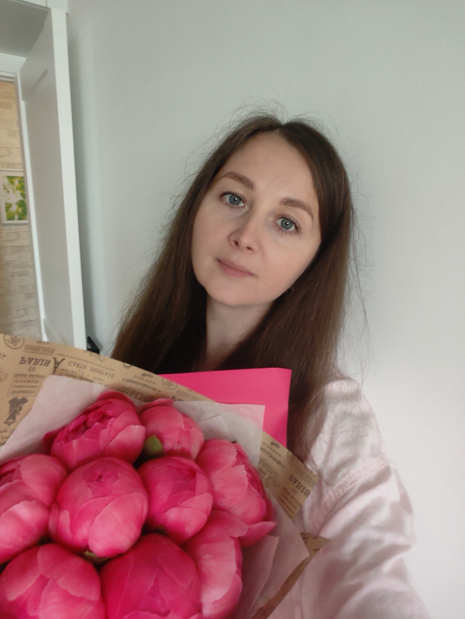 A medium shot captures a woman holding a vibrant bouquet of pink peonies and a bright pink card. The woman, with long brown hair and blue eyes, looks directly at the camera with a gentle expression. She is positioned slightly off-center to the right, against a plain light-colored wall. Her attire appears to be a light-colored, possibly pink, garment with a collar.
The bouquet of peonies, consisting of many tightly clustered, brilliant pink blooms, occupies the lower left portion of the frame. The flowers are wrapped in light brown paper that features faint, stylized patterns and inverted text, which appears to be in Cyrillic script. Partially visible words include "БУКЕТ" (BOUQUET) and what might be "CYALE МОЛЛИТЕ" (possibly part of a phrase like "SOUVENIRS PRAY" or a brand name). A distinct bright pink card or envelope is tucked into the wrapping, peeking out from behind the flowers.
In the background, to the left, a sliver of a white door frame is visible next to a light brick-patterned wall. A small framed picture, showing green and white abstract shapes, hangs on this wall. The setting appears to be indoors, likely a home, with soft ambient lighting. The overall mood conveyed is calm and appreciative.