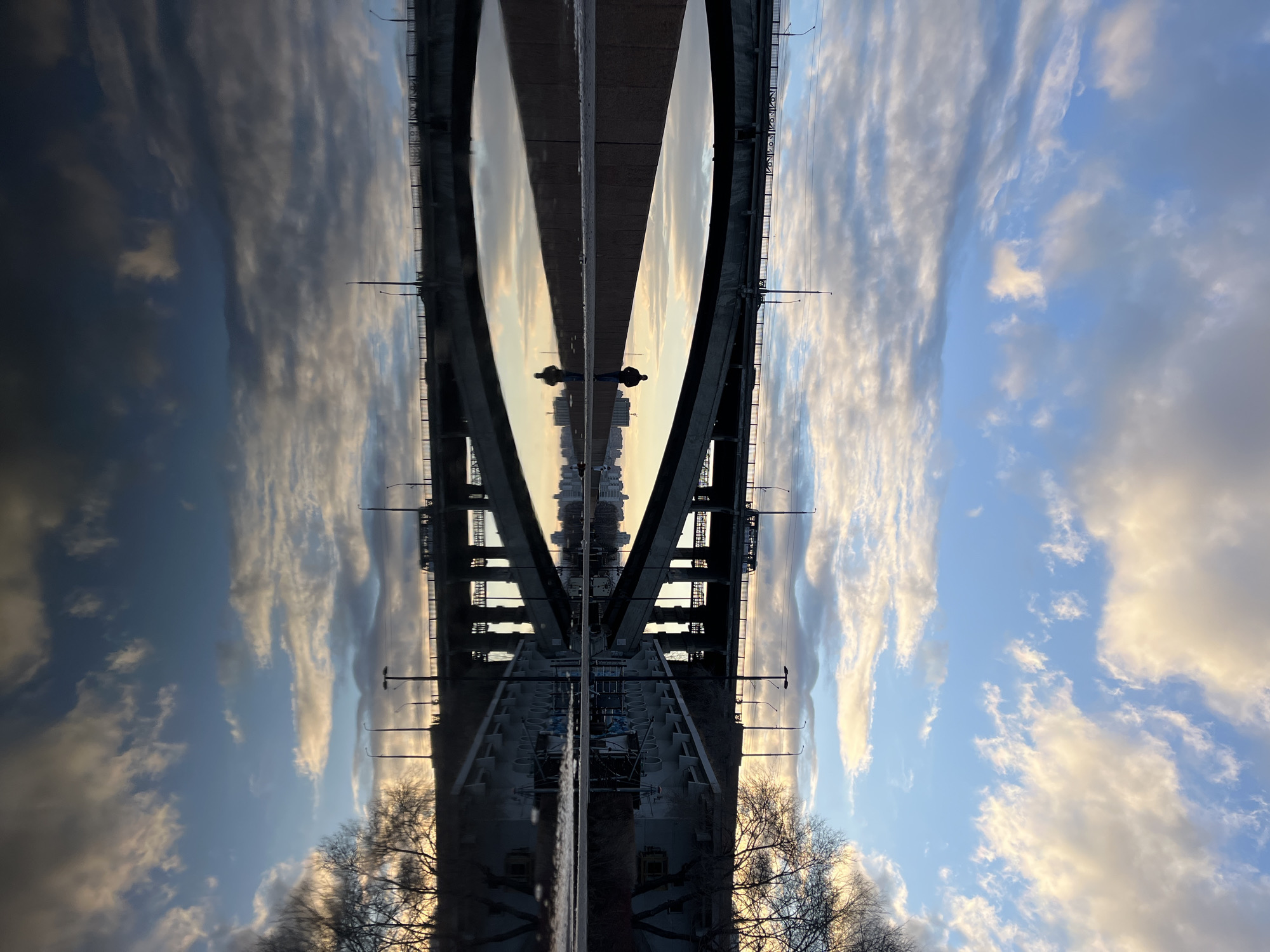 This image presents a striking, vertically symmetrical composition of a bridge reflected in a body of water, creating an optical illusion. The scene is set in an urban or semi-urban environment, likely a river in Krasnoyarsk, Russia.
The upper half of the image displays the actual scene: a wide bridge with a visible road surface, railings, and lampposts. The bridge's structure appears to be made of concrete or metal. Beyond the bridge, in the distance, faint outlines of city buildings are visible against the horizon. The sky above is partly cloudy, featuring soft blues, whites, and warm tones of yellow and orange, indicative of either sunrise or sunset. On the lower edges of this upper half, silhouetted bare tree branches can be seen, suggesting late autumn or winter.
The lower half of the image is a near-perfect, inverted reflection of the upper scene, cast upon a calm, highly reflective body of water, presumably a river or large lake beneath the bridge. The sky's colors and cloud patterns are mirrored, as are the bridge's structural elements, lampposts, and distant city skyline. Two small, dark, human figures are visible on the bridge's road surface, and their inverted reflections appear in the water below them. The bare trees seen in the upper half also have their reflections extending downwards.
The notable details include the soft, warm lighting of dawn or dusk, the varied cloud formations across the sky, and the crisp, clear reflection that creates the powerful visual symmetry. No discernible text is visible in the image. The overall impression is one of tranquility and visual harmony, with the reflection serving as a key element of the artistic composition.