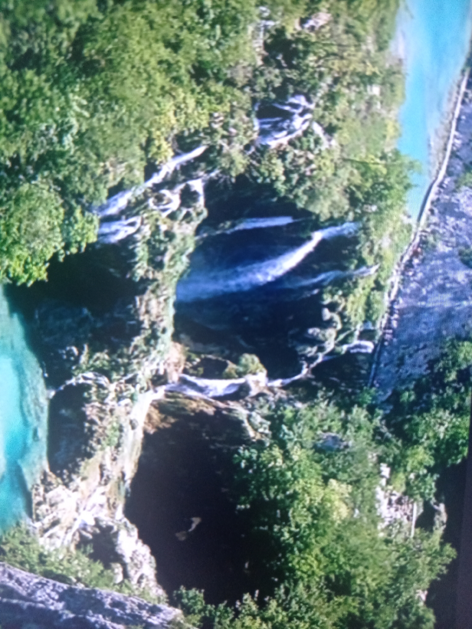 The image shows a top-down view of a natural landscape with lush green trees and water. There are waterfalls cascading down rocky cliffs into a clear, turquoise pool. The water appears shallow in some areas and deeper in others. The surrounding vegetation is dense and vibrant, suggesting a tropical or subtropical environment. On the right side of the image, there is a pathway or road made of stones, bordered by the vegetation and the water. The scene is well-lit, indicating it is likely daytime. The overall impression is one of natural beauty and tranquility. There are no visible people or objects, other than the natural elements. There is no visible text in the image.