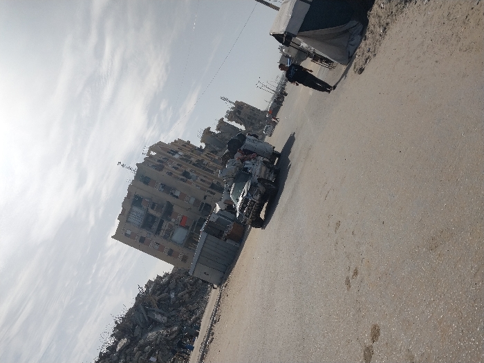 The scene in Khan Yunis, Palestinian Territory, depicts a damaged urban environment under a partly cloudy sky. An asphalt road spans the foreground and mid-ground. To the left, a substantial pile of building rubble, composed of concrete and twisted rebar, sits adjacent to a multi-story building exhibiting significant structural damage, including shattered windows and exposed interiors. Two small, gray metal kiosks or temporary structures are situated near the base of the damaged building. A light-colored pickup truck, visibly damaged on its front and hood, is positioned diagonally on the road. At least three individuals are observed in or near the truck's bed, one appearing to wear a keffiyeh. On the right side of the road, a single male figure in dark clothing walks away from the viewer, passing a makeshift tent erected on sandy ground adjacent to the road.