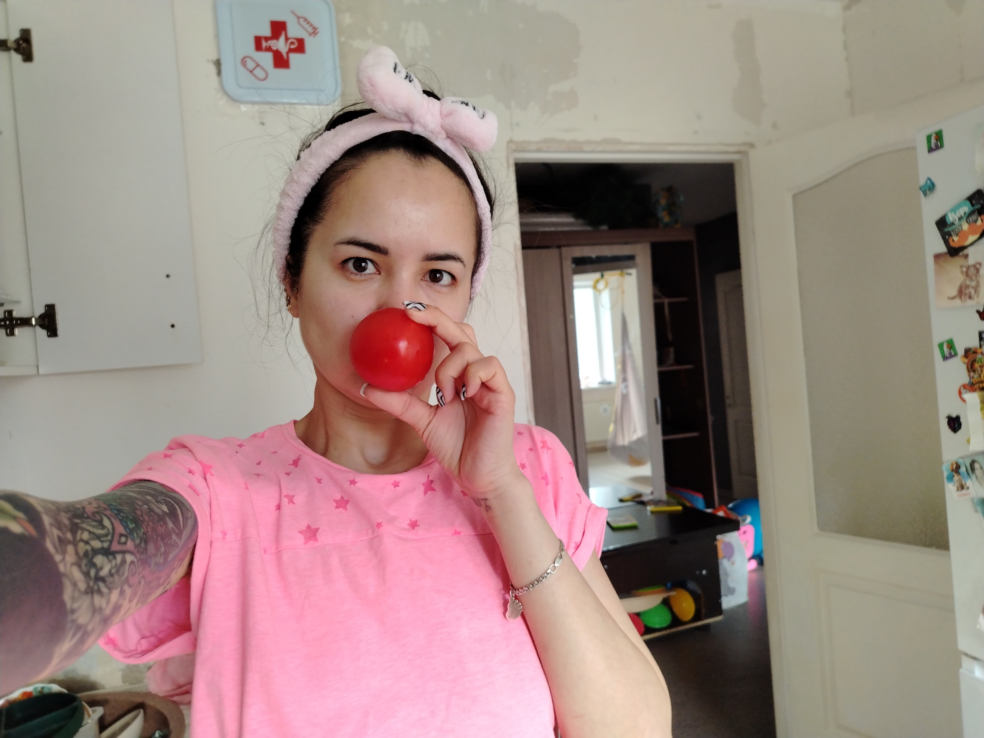 A woman with a pink headband with a bow on top is holding a red tomato to her nose. She is wearing a pink t-shirt with stars on it and has a tattoo on her left arm and a bracelet on her right wrist. The woman is in a room with a kitchen cabinet on the left and a refrigerator on the right. In the background is a doorway leading to another room with a table and cabinet. A blue sign with medical symbols is on the wall behind the woman. The location is Noviy, Russia.