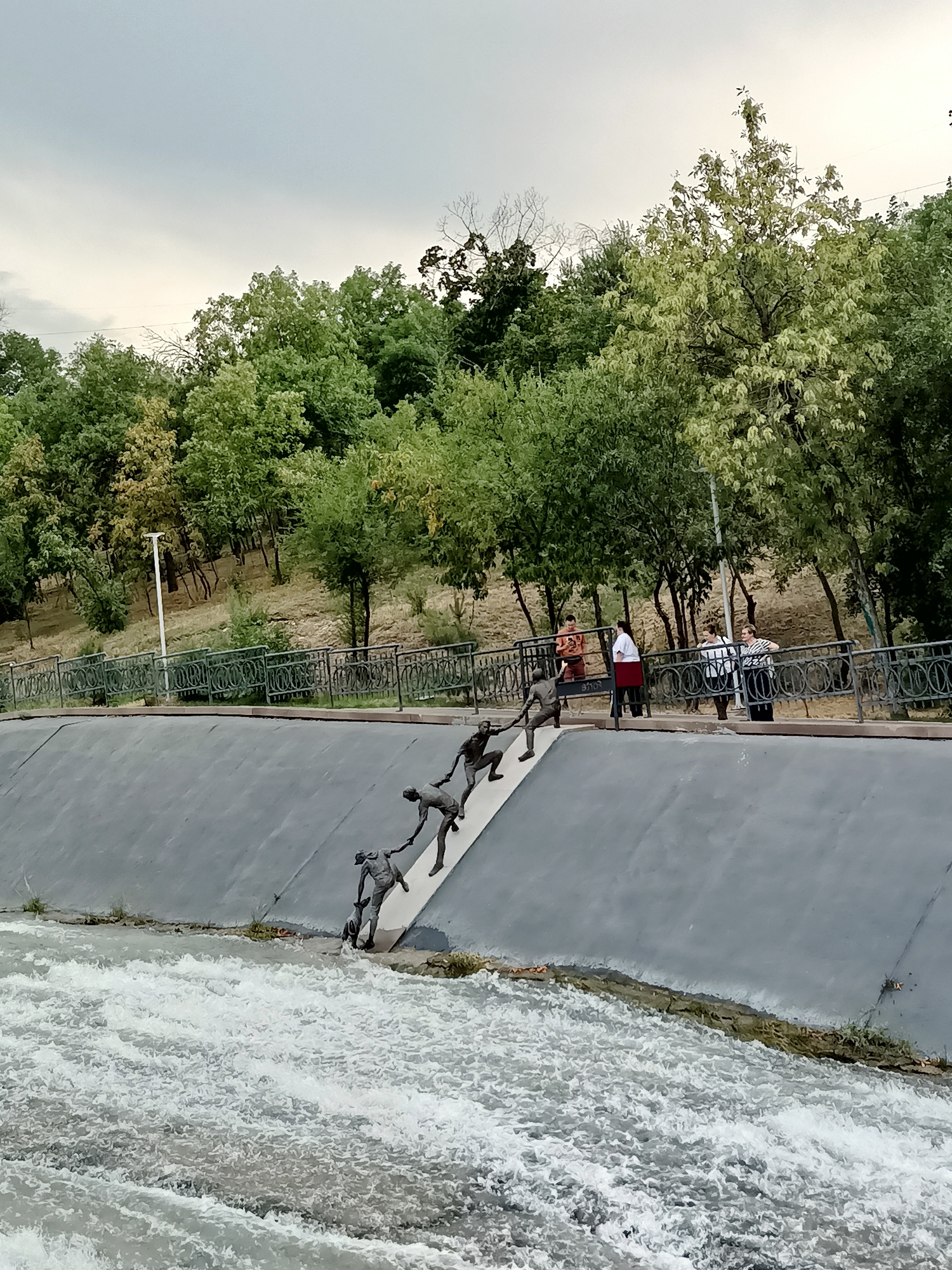 In Almaty, Kazakhstan, a sculpture depicting several figures struggling to ascend a steep incline is visible. The sculpture is situated on a grey concrete embankment adjacent to turbulent water. Above the embankment, a metal fence separates the area from a park with trees and a pathway where four individuals stand observing the sculpture. The sky is overcast.