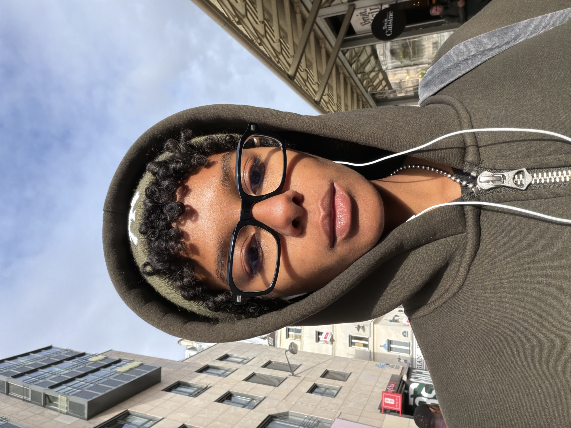 The image is a selfie taken at an upward angle in Paris, France. The subject is wearing glasses, a hoodie, and headphones. Buildings are visible in the background, including a modern building with multiple windows and a traditional Parisian building with window boxes. A sign for "Petit Saint Cuisine" is visible in the upper right corner. The sky is visible above the buildings. The subject appears to be standing on the street, looking up.
