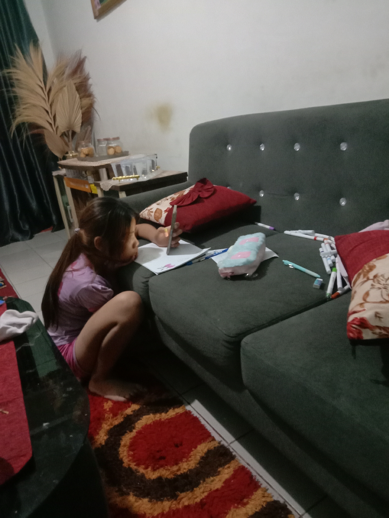 A young girl with her hair in a ponytail, wearing a pink t-shirt and shorts, is sitting on the floor next to a dark green couch in a dimly lit room. She is looking at a piece of paper and holding a pen. Several markers and a pencil case are scattered on the couch. A red pillow with a dark bow is on the couch behind her. A patterned rug is on the floor in front of the couch. In the background, a small table with two glass jars containing cookies is visible, along with a dried palm leaf arrangement. The scene appears to be indoors, possibly a living room in Kwandang, Indonesia.
