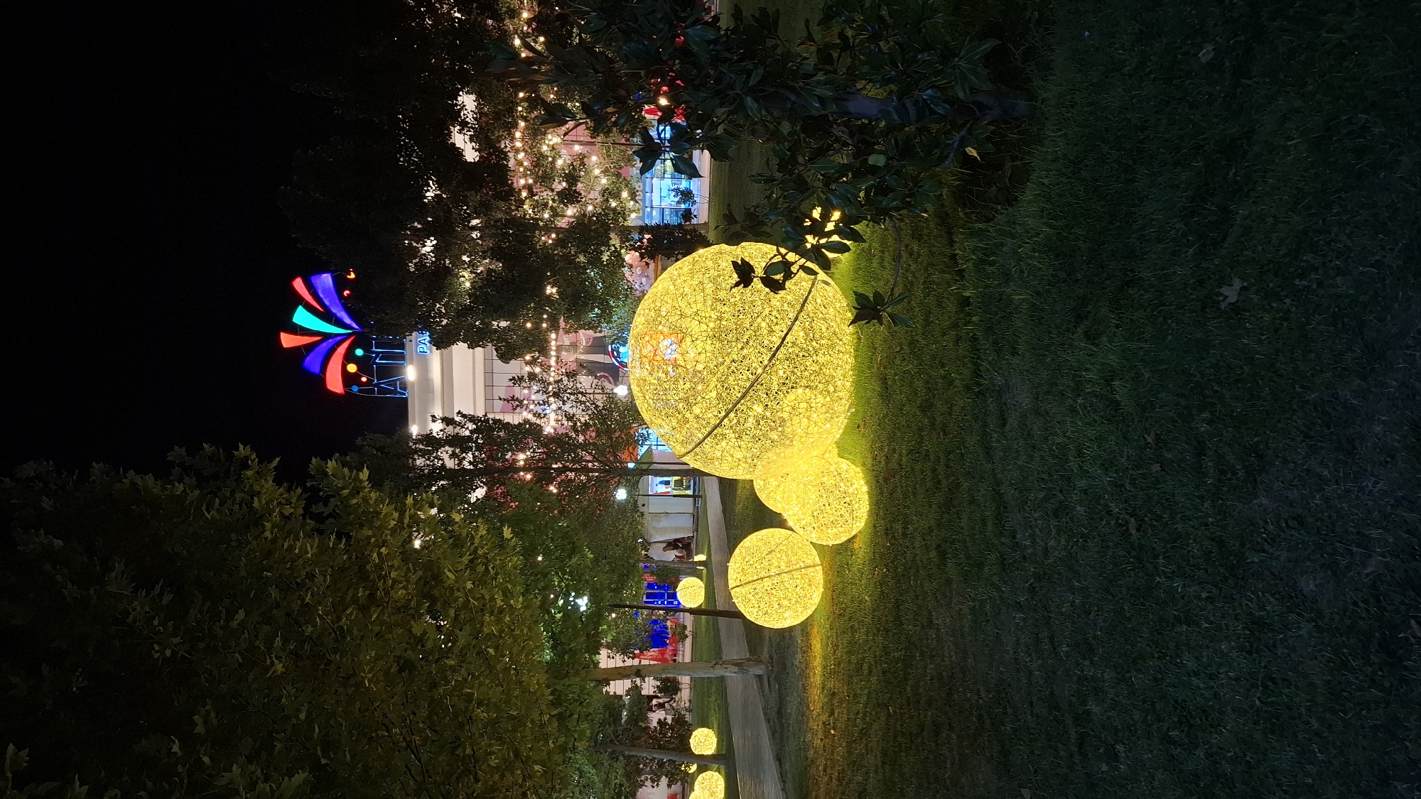 The image depicts an outdoor scene at night, likely a park or plaza, decorated with illuminated spherical light installations. In the foreground, several large, glowing yellow orbs are arranged on a grassy lawn. These orbs appear to be made of a woven material or mesh, with numerous small lights creating a radiant effect. Behind the orbs, a path or walkway is visible, lined with trees. Further in the background, a modern building with large windows is illuminated. Above the building, a colorful, fan-shaped light display is visible against the dark night sky. String lights are also visible in the trees. The overall atmosphere is festive and decorative, suggesting a public space designed for evening enjoyment or an event.