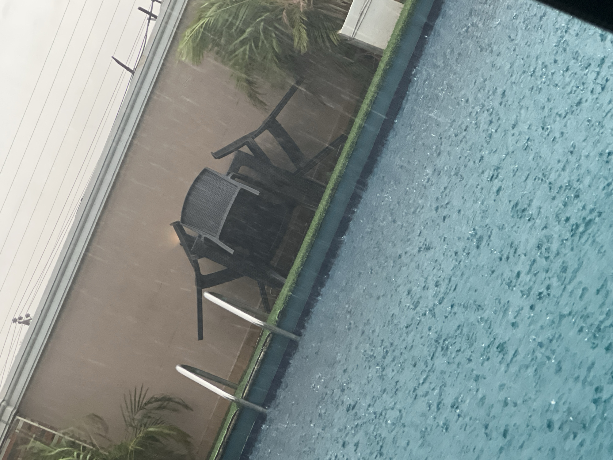 The image displays an outdoor swimming pool area captured during a period of heavy rainfall. The central subject is a light blue swimming pool, its surface densely covered with numerous ripples and splashes caused by the falling raindrops, clearly indicating a significant downpour. A metallic ladder with two visible rungs extends into the pool from the foreground.
Lining the edge of the pool is a strip of artificial green turf. Behind the pool, a light brown or beige wall runs horizontally across the frame. Further back and slightly obscured, green foliage, likely from palm trees, is visible, some leaves appearing blurred due to the rain or motion. Two dark-colored patio chairs, possibly made of wicker or a similar material, are positioned near the wall; one is partially visible on the left, and another appears tilted or stacked further to the right.
The sky in the upper portion of the image is overcast, suggesting daytime conditions despite the gloomy weather. Several parallel overhead wires or cables are visible stretching across the top left corner. The scene evokes the atmosphere of a tropical or subtropical climate, consistent with the provided location context of Abuja, Nigeria. No people are present in the image, and no visible text can be identified.