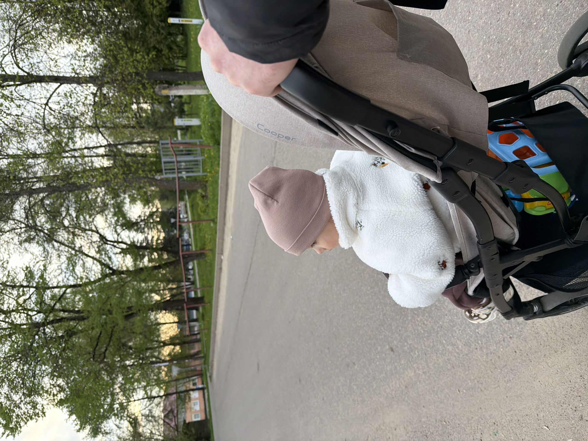 A toddler in a pink beanie and white teddy bear jacket sits in a "Cooper" stroller. The stroller is being pushed on a paved road in Reshetnikovo, Russia. Trees and some low-rise buildings are visible in the background. A person's hand holds the stroller handle. A colorful toy is stored in the stroller's storage basket.