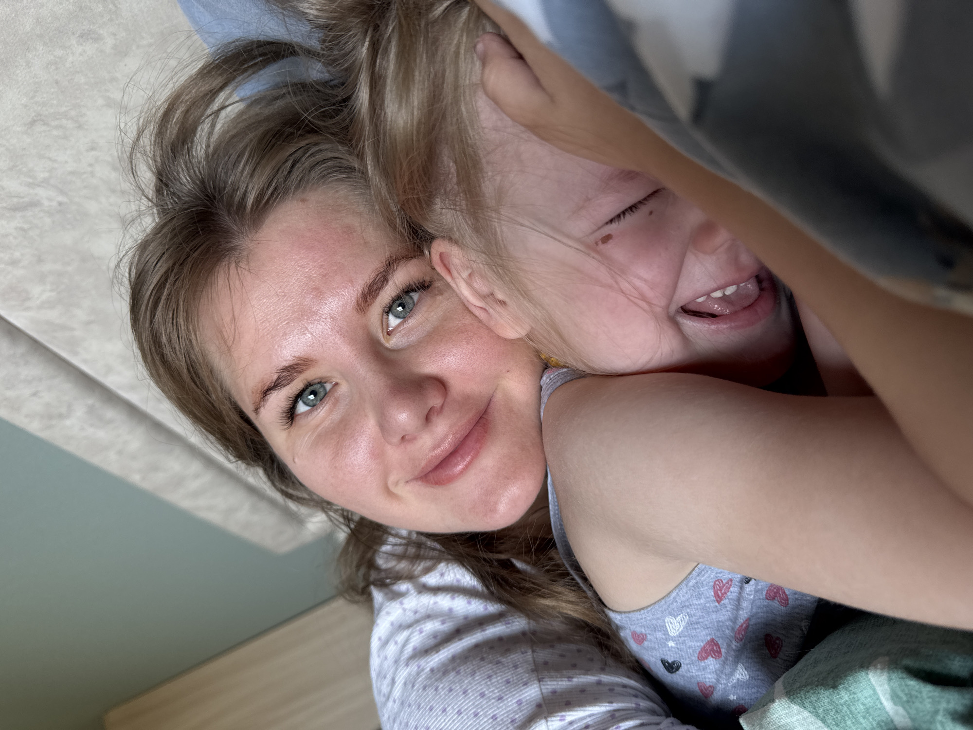 A close-up shot captures a woman with light skin, light brown hair, and striking light blue eyes, embracing a young child. The child, also fair-skinned with blonde hair, is laughing with their eyes closed. The woman is wearing a white top patterned with small grey dots. The child is dressed in a grey tank top adorned with heart shapes. The background is blurred, showing a light green wall and a wooden surface. The location is presumed to be indoors, possibly in a home in Ферма, Russia, though this cannot be confirmed from the image. The interaction between the two subjects is affectionate.