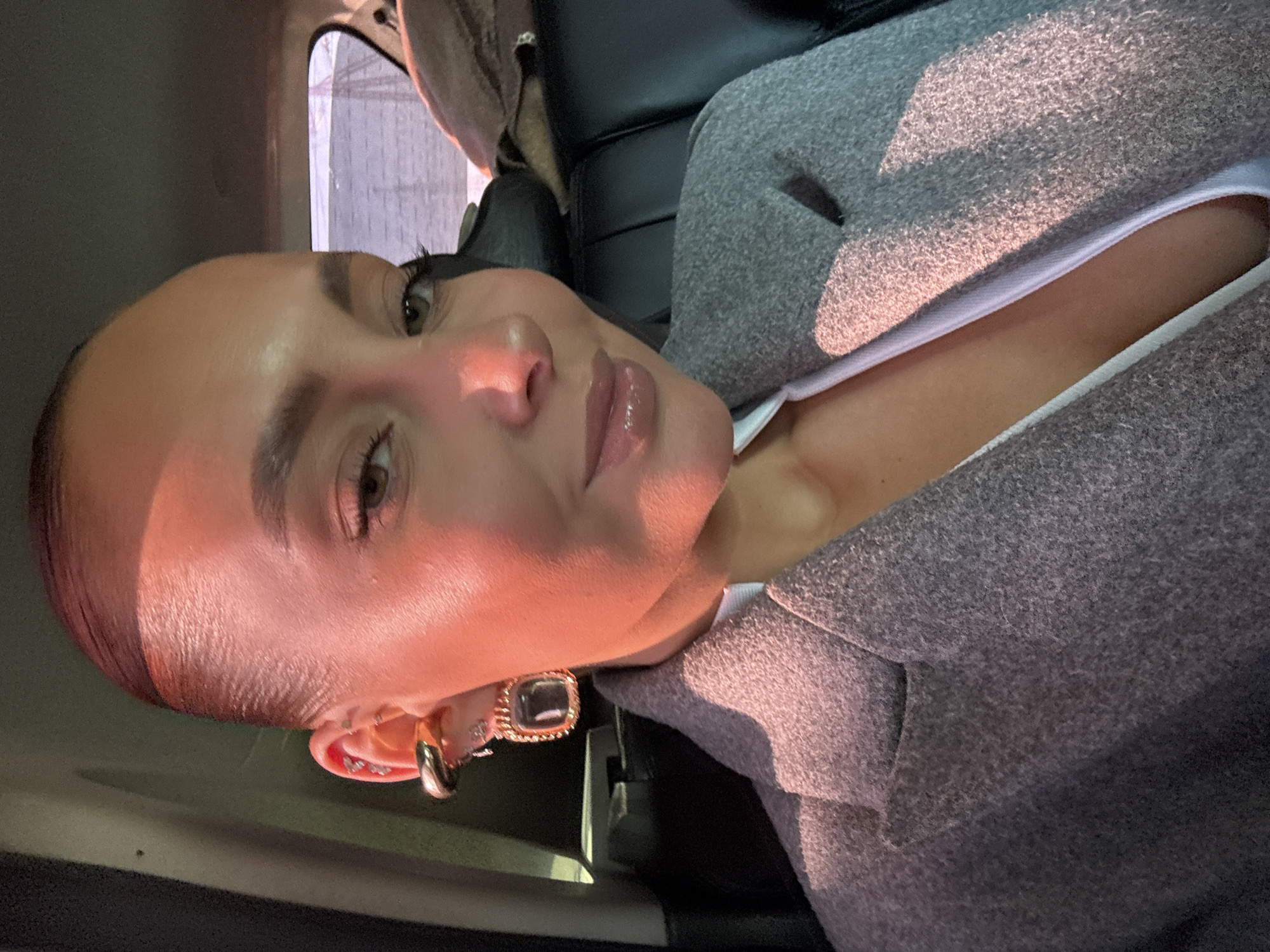 An adult female is captured in a close-up selfie, seated within the passenger compartment of a vehicle. Her head and upper torso are visible, positioned centrally in the frame. She has dark hair pulled back, light brown eyes, sculpted eyebrows, and full lips accentuated with gloss, exhibiting a slight smile directed at the camera. Her visible left ear features multiple adornments, including a large, polished gold-toned ear cuff on the upper helix, a prominent square-shaped gold-framed earring with a dark, possibly transparent, insert dangling from the earlobe, and several smaller metallic studs along the helix. She wears a grey, textured blazer or coat with a notched lapel, layered over a white garment, the neckline of which is partially visible. Intense, warm-toned lighting, indicative of direct sunlight or a strong artificial source, illuminates her face from the right, casting distinct highlights on her forehead, cheekbone, and nose, imparting a reddish hue to her skin. The vehicle's interior features dark, likely leather, seating and a dark fabric headliner. Through the rear window, a blurred outdoor landscape is visible, suggesting a body of water under a light sky, with faint, possibly structural, outlines or power lines on the distant horizon. This scene is situated within the city of Chaikovsky, Russia, though the precise location within the city is not identifiable from the image.