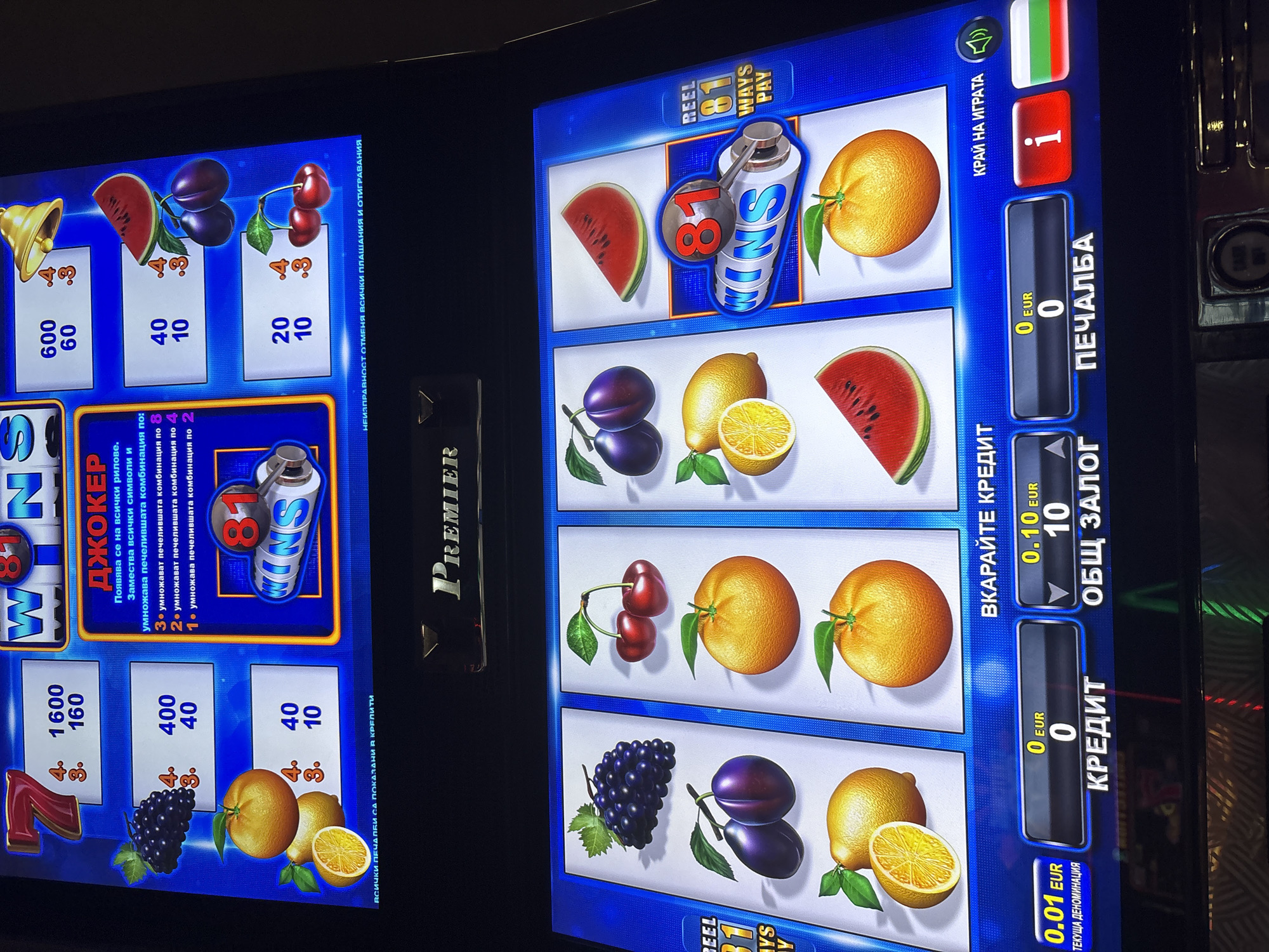 The image shows a close-up of a slot machine in Karlovo, Bulgaria. The machine displays various fruit symbols, including grapes, plums, oranges, lemons, and watermelons, as well as numbers and text in Bulgarian. The top part of the screen shows winning combinations and payouts, with "ДЖОКЕР" (Joker) highlighted. The bottom part of the screen displays betting information, with "КРЕДИТ" (Credit), "ОБЩ ЗАЛОГ" (Total Bet), and "ПЕЧАЛБА" (Winnings) shown in Euros. A Bulgarian flag is visible on the right side of the screen. The machine is lit up and appears to be in operation. The setting is likely an indoor entertainment venue, such as a casino or a bar. There are no people visible in the image.
