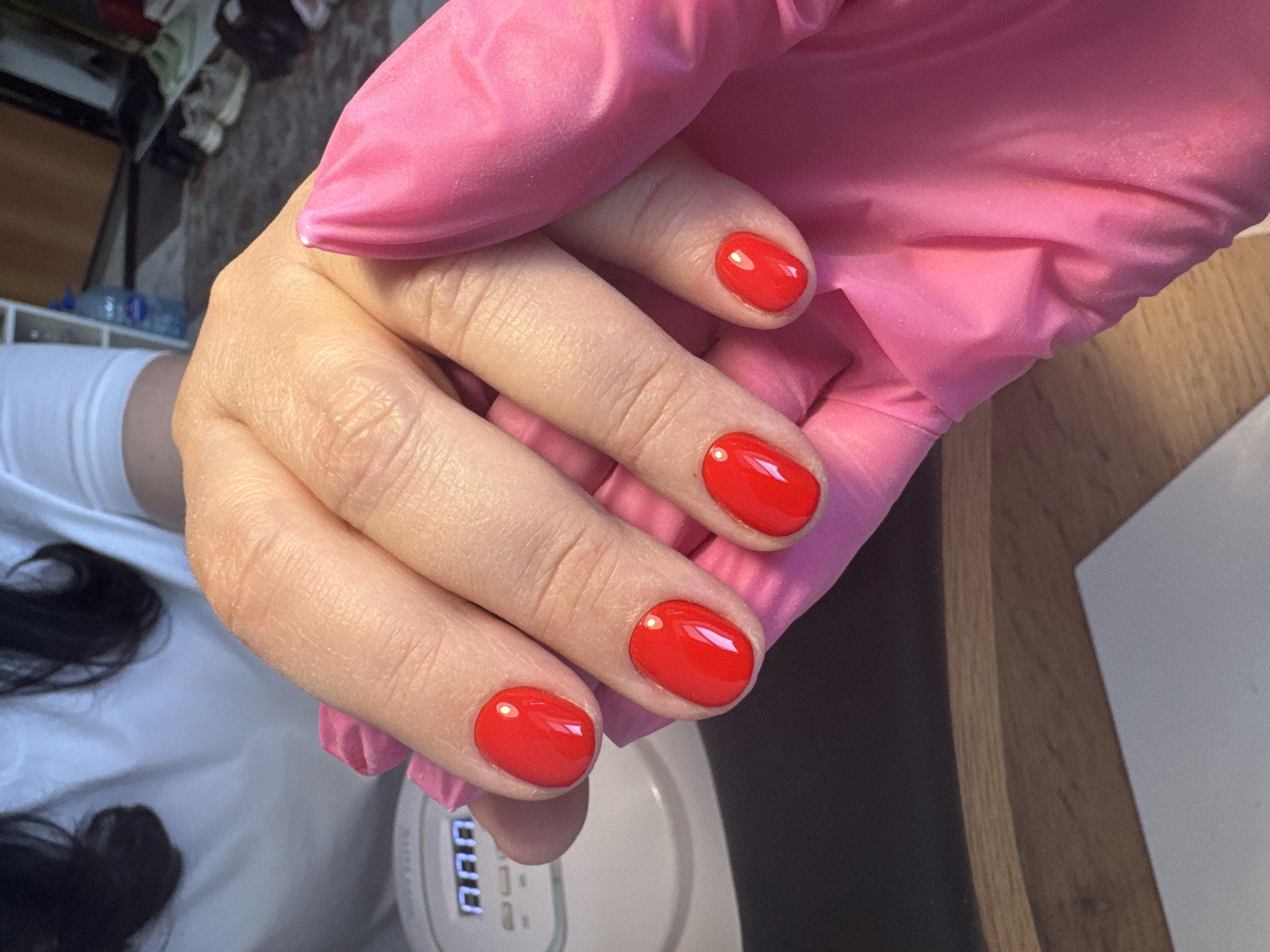 The image shows a person's left hand, wearing a pink rubber glove, positioned above a white UV nail lamp. The fingernails of the hand are painted with a glossy red polish. The background includes glimpses of a white surface, dark hair, and a wooden surface. The image was taken indoors. The location is not discernible from the image.