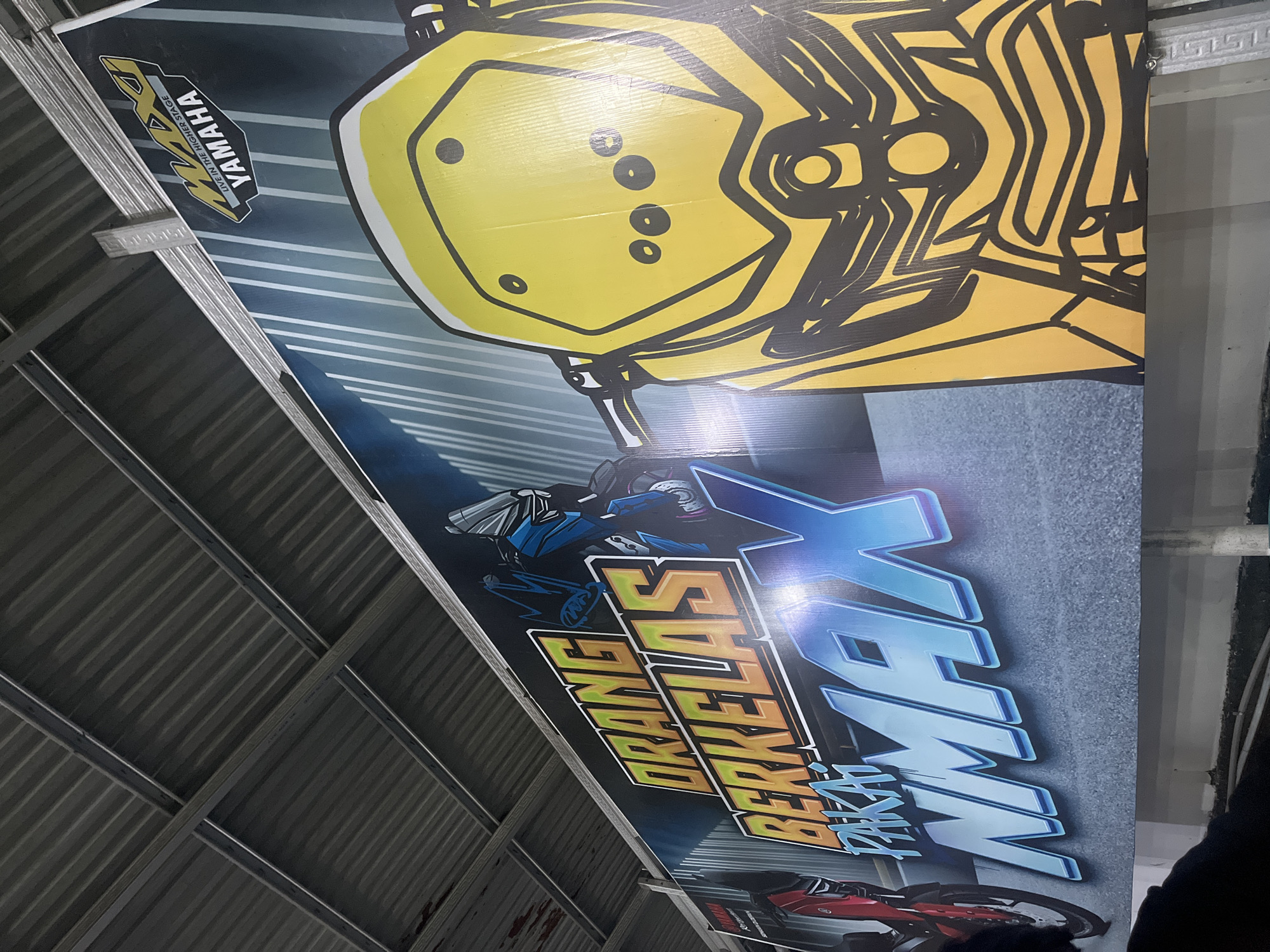 The image shows a large banner advertising Yamaha NMAX motorcycles. The banner has text that reads "ORANG BERKELAS PAKAI NMAX" which translates to "Classy people use NMAX". There is also a Yamaha logo on the banner. The banner is displayed outdoors under a corrugated metal roof, suggesting it is in a covered area, possibly a dealership or a public space. The style of the roof and the overall aesthetic do not strongly indicate a specific location within Banda Aceh, Indonesia, but the presence of the banner suggests it is in an urban or semi-urban setting where such advertisements are common. The banner features stylized illustrations of motorcycles, with a prominent yellow NMAX front fairing on the right side and parts of red and blue motorcycles on the left. The lighting suggests it may be daytime, but the reflection on the banner indicates artificial lighting might also be present. No people are clearly visible interacting with the banner.