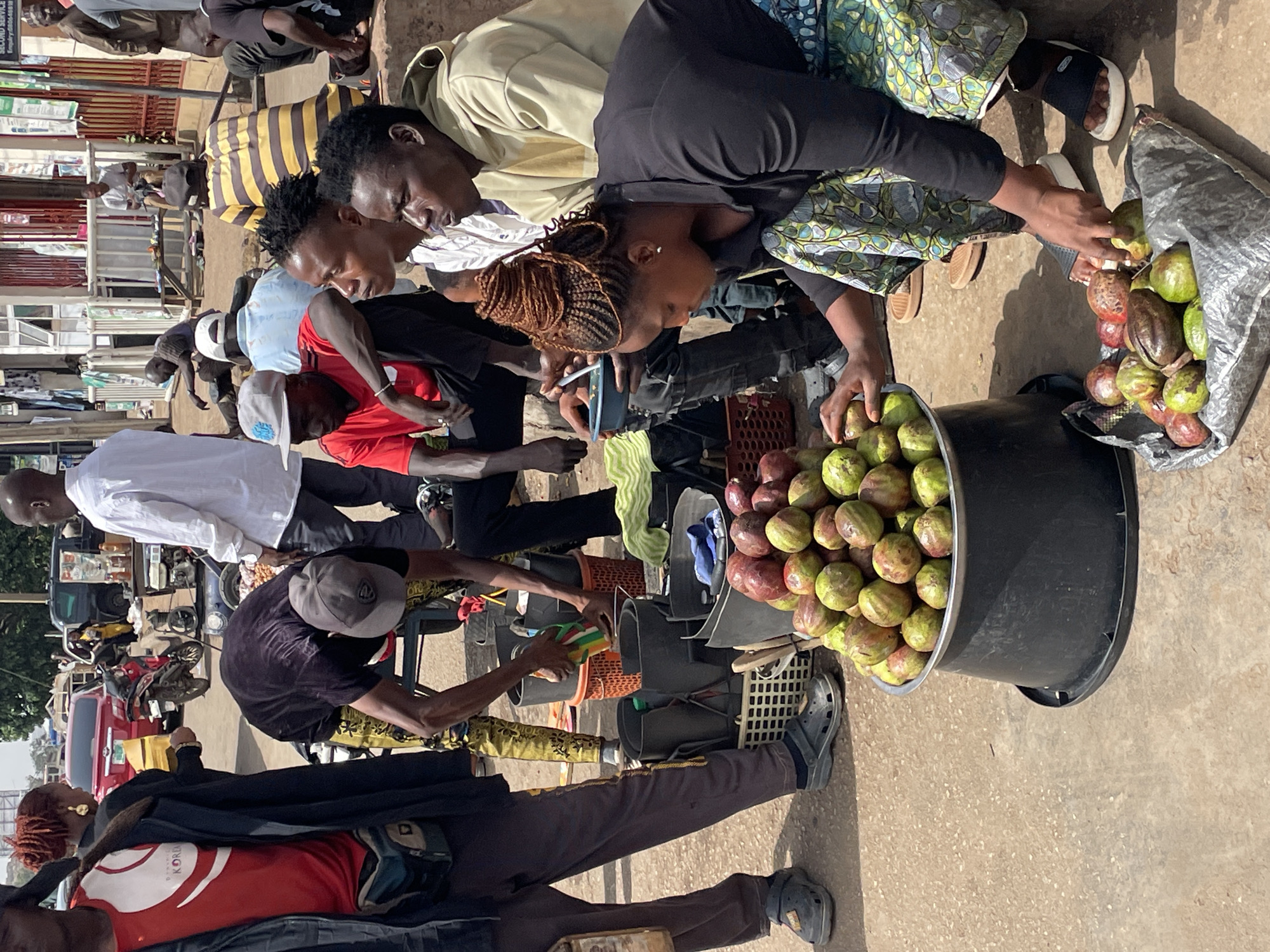 The image captures a busy street scene in Jos, Nigeria, focused on a fruit vending operation. Several individuals are clustered around a mobile vending setup that consists of a large black bucket filled with round, reddish-green fruits, likely African pears or avocados. A woman in a patterned dress is transferring fruit from a plastic bag into the bucket. Other people are seen examining or preparing to purchase the fruits. The foreground features legs and lower torsos of people walking past. The background shows urban elements like buildings, motorcycles, and parked vehicles. The time of day appears to be mid-morning or early afternoon due to the angle of the sunlight.