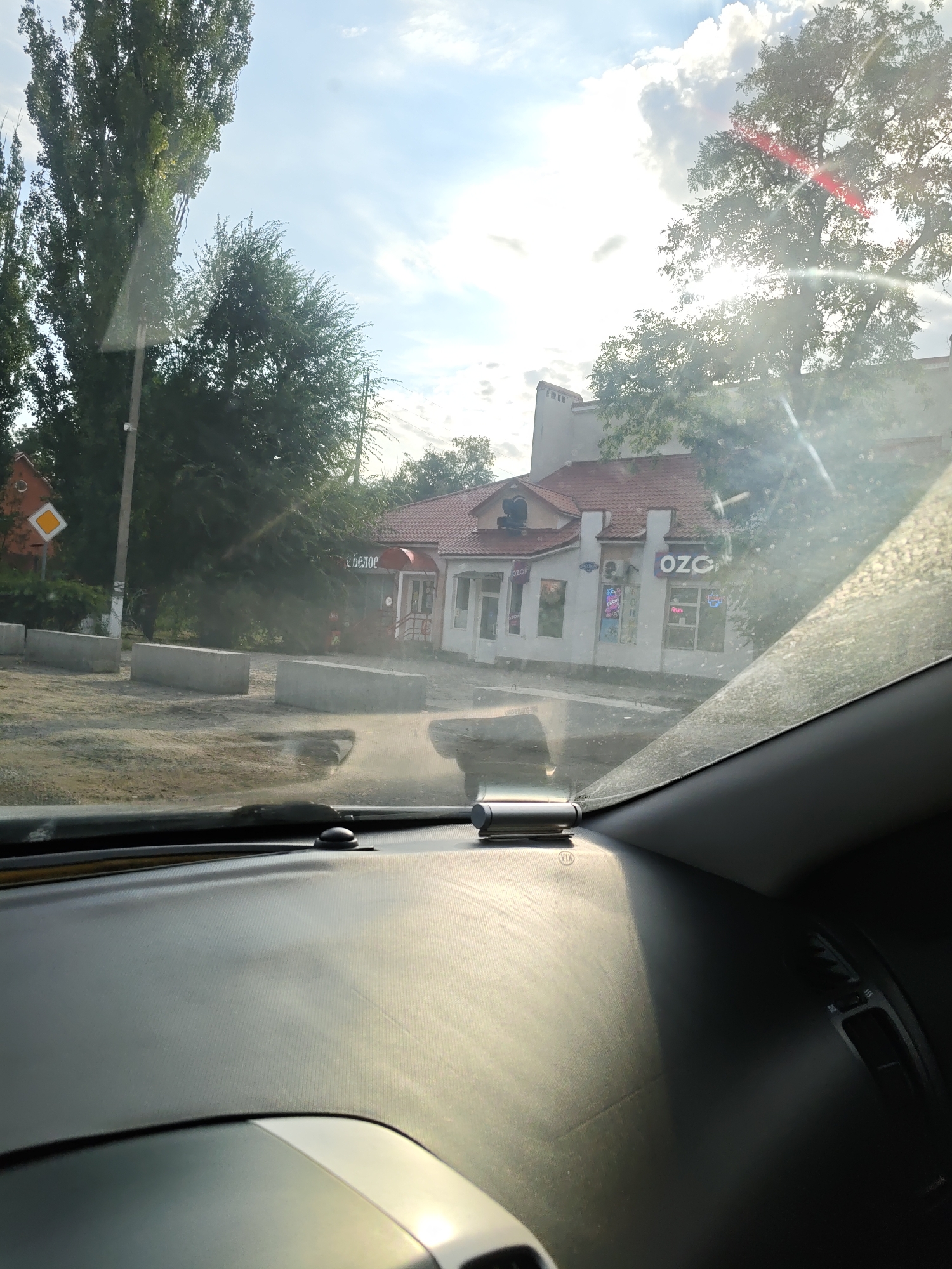 The image shows a view from inside a vehicle, looking out at a street scene. In the foreground, the dashboard of the car is visible, with a car mount for a device. The windshield is dusty and reflects sunlight.
Outside the car, there is a street with a dirt and gravel surface, possibly undergoing construction, as evidenced by several concrete blocks lined up. A traffic sign is visible on the left.
Further back, a building with a red tiled roof is situated. It appears to be a commercial establishment with signage that includes "OZO". There are trees and power lines in the background, and the sky is partly cloudy with bright sunlight. The location is identified as Persianovskiy, Russia. No people are visible in the image.