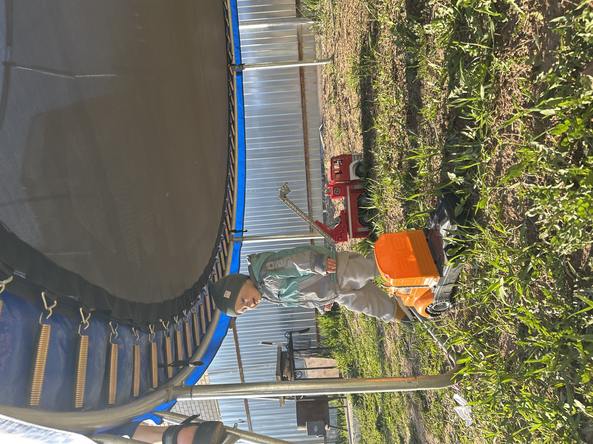 A young child wearing a dark beanie, gray jacket, and gray pants is standing next to a large orange toy truck on grass. The scene is outdoors, with a trampoline visible to the left and a metal fence in the background. A red toy fire truck is also visible. The location is in the city of Chaikovsky, Russia.