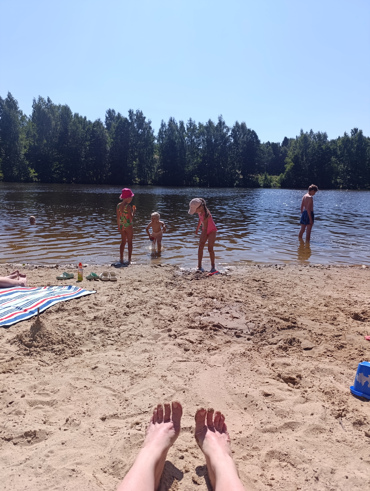 A group of people are at a beach on a sunny day. In the foreground, a person's feet are visible on the sand. A striped towel is laid out on the sand next to a small sandcastle. Scattered around are several pairs of sandals and a bottle of sunscreen. Further back, four children are in the shallow water. One child, wearing a pink hat and swimsuit, stands near the shore. Another child, wearing a pink swimsuit and a white hat, is bent over. A third child is partially submerged in the water, and a fourth child, wearing blue swim trunks, stands further out. The water is calm and reflects the clear blue sky. The background is lined with dense green trees.