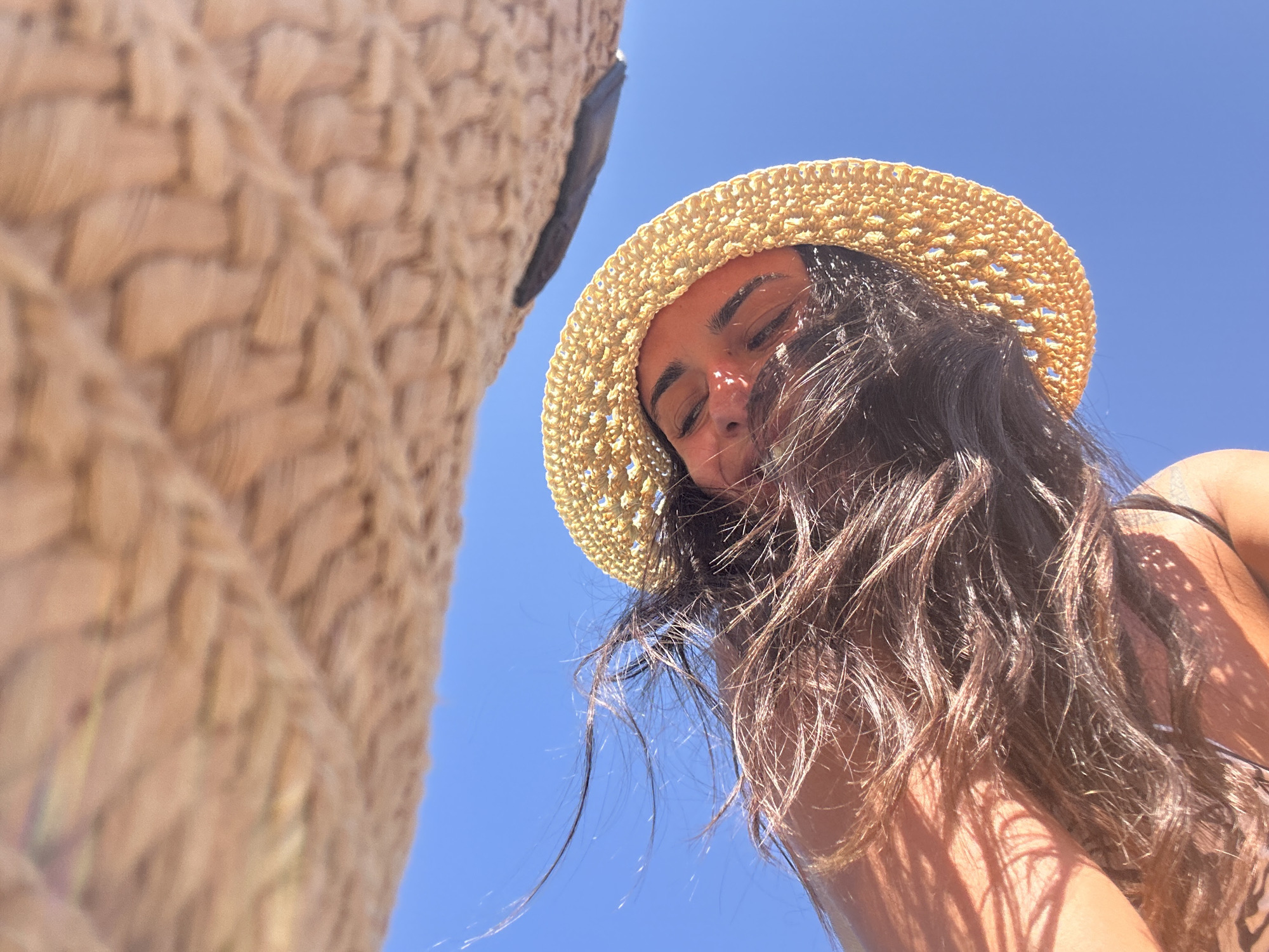 An overhead shot shows a person wearing a straw hat and looking up at the sky in Cala Lliteres, Spain. The person's hair is blowing in the wind, and shadows from an overhead object, likely a sun umbrella, are cast across their face. The sky is clear and blue, indicating a sunny day.
