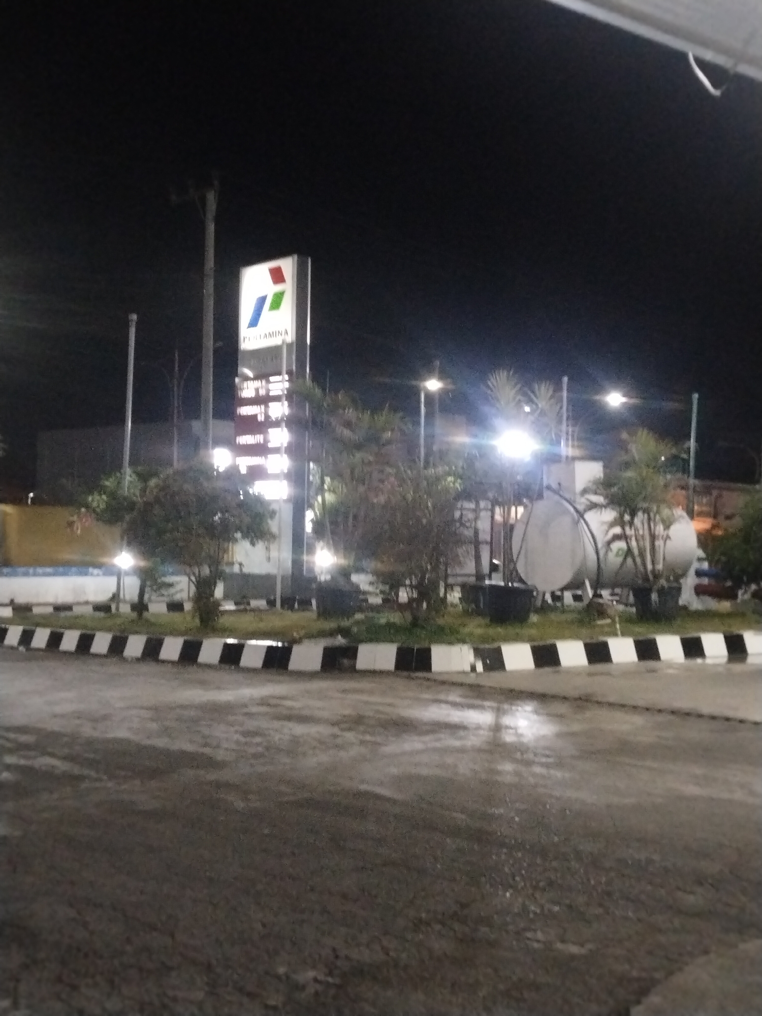 This image captures a nighttime scene at a Pertamina gas station in Keuniree, Indonesia. A prominent, illuminated vertical sign stands in the mid-ground, featuring the multi-colored Pertamina logo (red, green, blue elements) at the top, followed by the "PERTAMINA" text and a list of fuel types such as "PERTALITE" and "PERTAMAX" with their corresponding prices, though specific values are unreadable due to distance and lighting. The ground surface, likely asphalt or concrete, is visibly wet in various sections, reflecting ambient light. The station forecourt includes landscaped islands bordered by distinct black and white striped curbs, containing several small trees and shrubs. Behind the vegetation on the right side of the frame, two large, horizontally oriented, cylindrical white storage tanks are visible. Multiple streetlights or station lights provide artificial illumination, creating bright spots and glare. Utility poles are present in the background. No people or vehicles are visible in the image, and no specific actions or interactions are depicted.