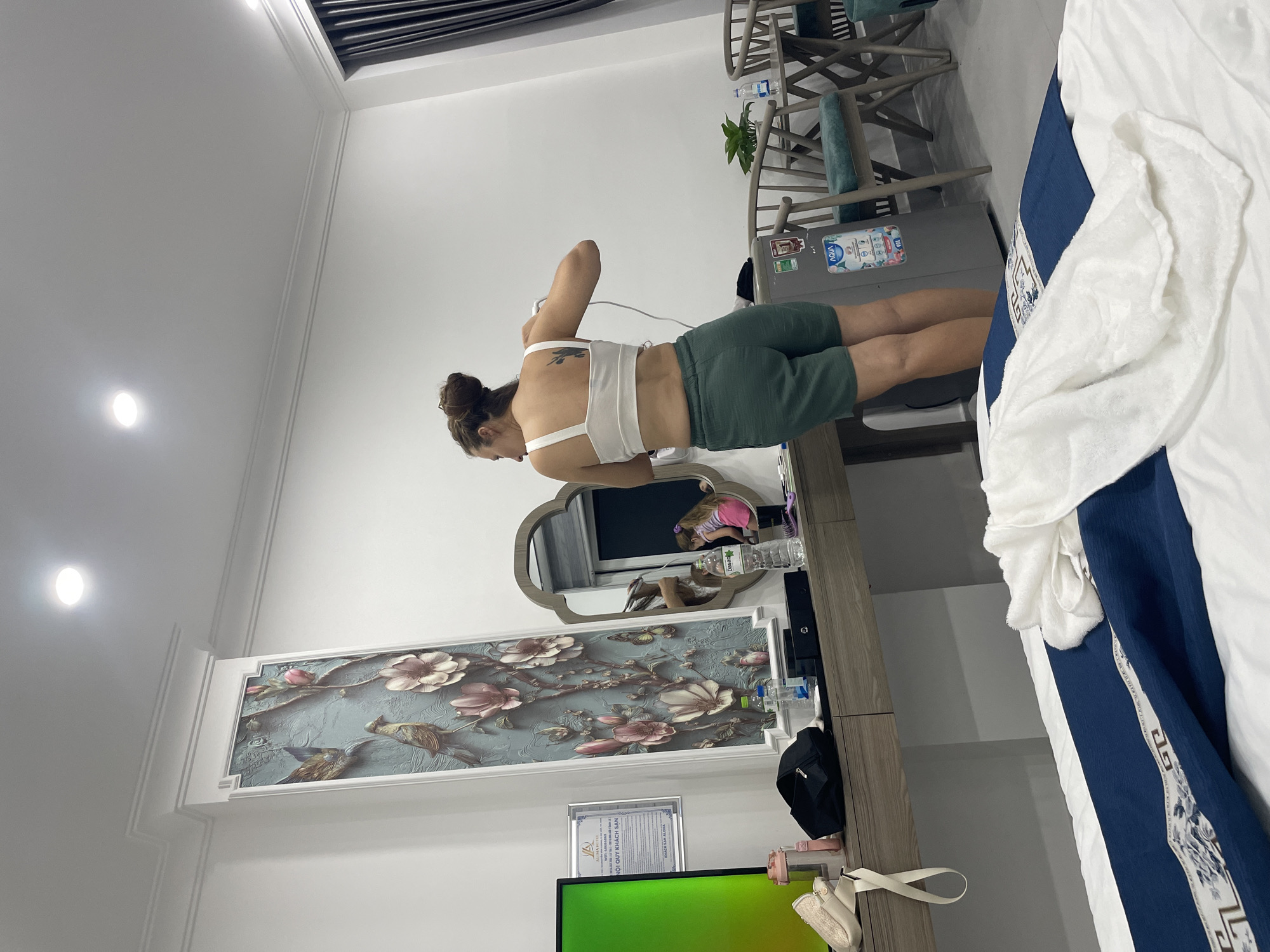 A person is standing in a room, facing away from the camera and towards a dresser with a mirror. The person is wearing a white tank top and green shorts. They have their hair pulled up in a bun and a tattoo on their back. The dresser is cluttered with various items, including bottles, makeup, and a small black bag. Behind the dresser, there is a refrigerator with stickers on it, and beyond that, several chairs and a plant. The room appears to be a bedroom or a dressing area, with a large bed visible on the right side of the image, covered in blue and white bedding. On the wall, there is a decorative panel featuring birds and flowers, and a framed certificate is visible below it. The lighting suggests it is daytime. The overall impression is a casual, lived-in space. The text "AGUA" and "DƯỢC PHẨM" are visible on labels on the refrigerator. A framed certificate contains Vietnamese text.