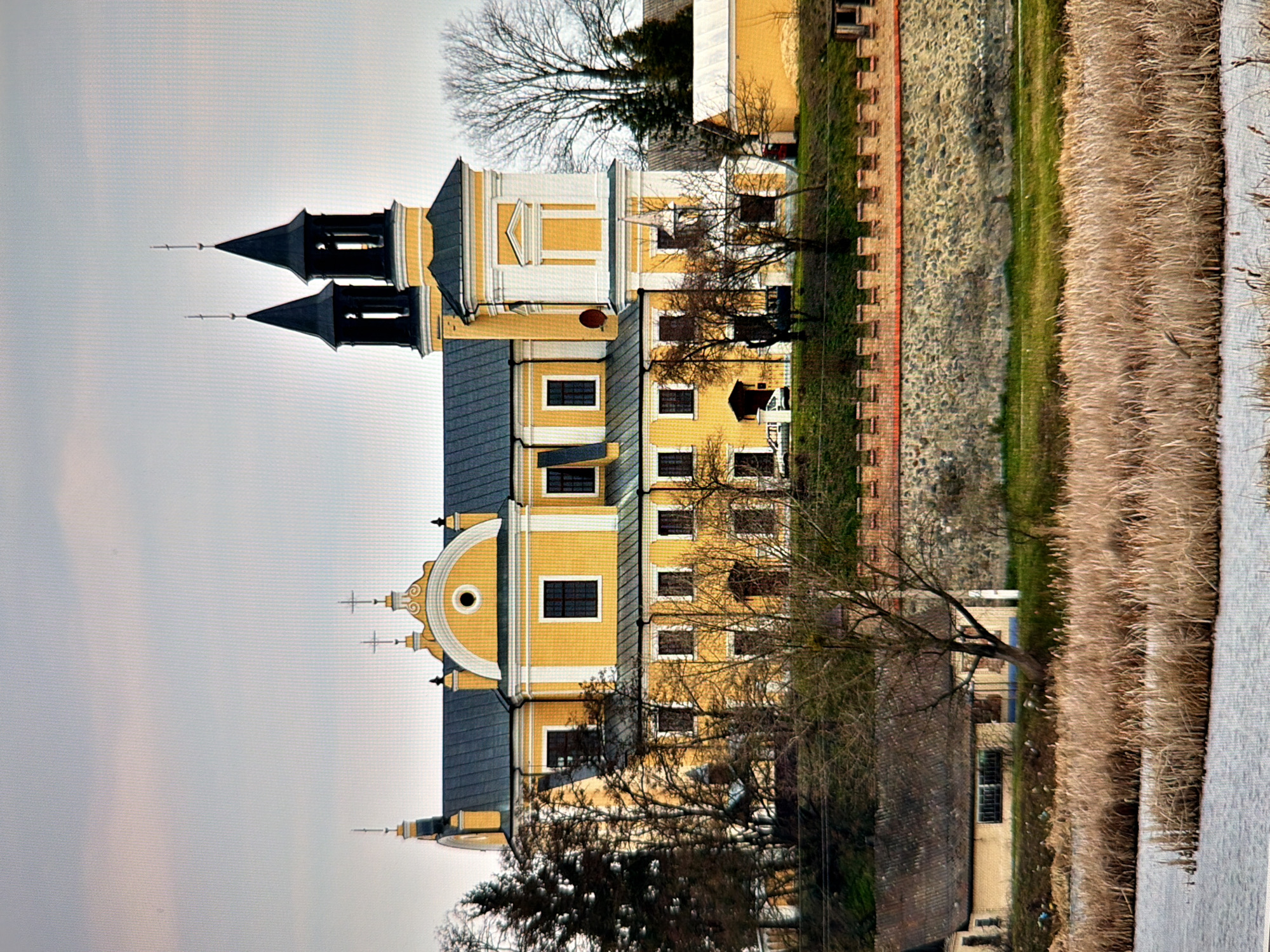 This is a landscape photograph of a large, yellow, Baroque-style building, likely a monastery or church, with two prominent dark spires topped with crosses. The building features white architectural details and a dark gray roof. In front of the main building, there is a lower yellow structure with a pitched roof. The building is situated on a grassy hill behind a stone and brick wall. To the left of the main building, there are several tall, bare trees. In the foreground, there is a body of water with dry reeds growing along its edge. The sky is overcast and light gray with hints of pink. The scene appears to be a rural or semi-rural setting, possibly on the outskirts of Kyiv, Ukraine. There are no people visible in the image.