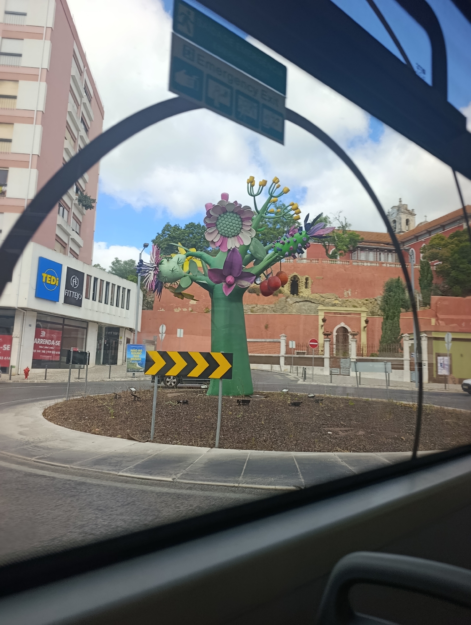 The image is a street-level view captured from inside a vehicle, looking out through the windshield. The focus is on a roundabout in the city of Setúbal, Portugal, decorated with a large, colorful, abstract sculpture resembling a tree with oversized flowers and fruit. To the left of the roundabout, a pink building with many windows and shutters stands tall. Below this building, storefronts are visible, including one with a blue "TEDI" sign and another with "FITTEJO" signage. A red fire hydrant is present on the sidewalk. In the foreground, a road curves around the roundabout, with a yellow and black chevron traffic sign indicating a turn. The background features a red-walled building with arched doorways and windows, possibly a historical structure, and a church steeple is visible above it. The sky is partly cloudy, with patches of blue.
