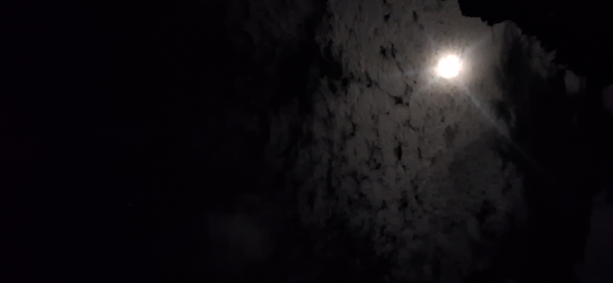 The image depicts a night scene in Vallecito, Venezuela. The right side of the frame is dominated by illuminated clouds, with a bright moon visible through them. The left side of the image is dark, with only a faint suggestion of clouds or other features. There are no visible people or objects. The scene suggests a dimly lit night, likely illuminated by the moon reflecting off the clouds.