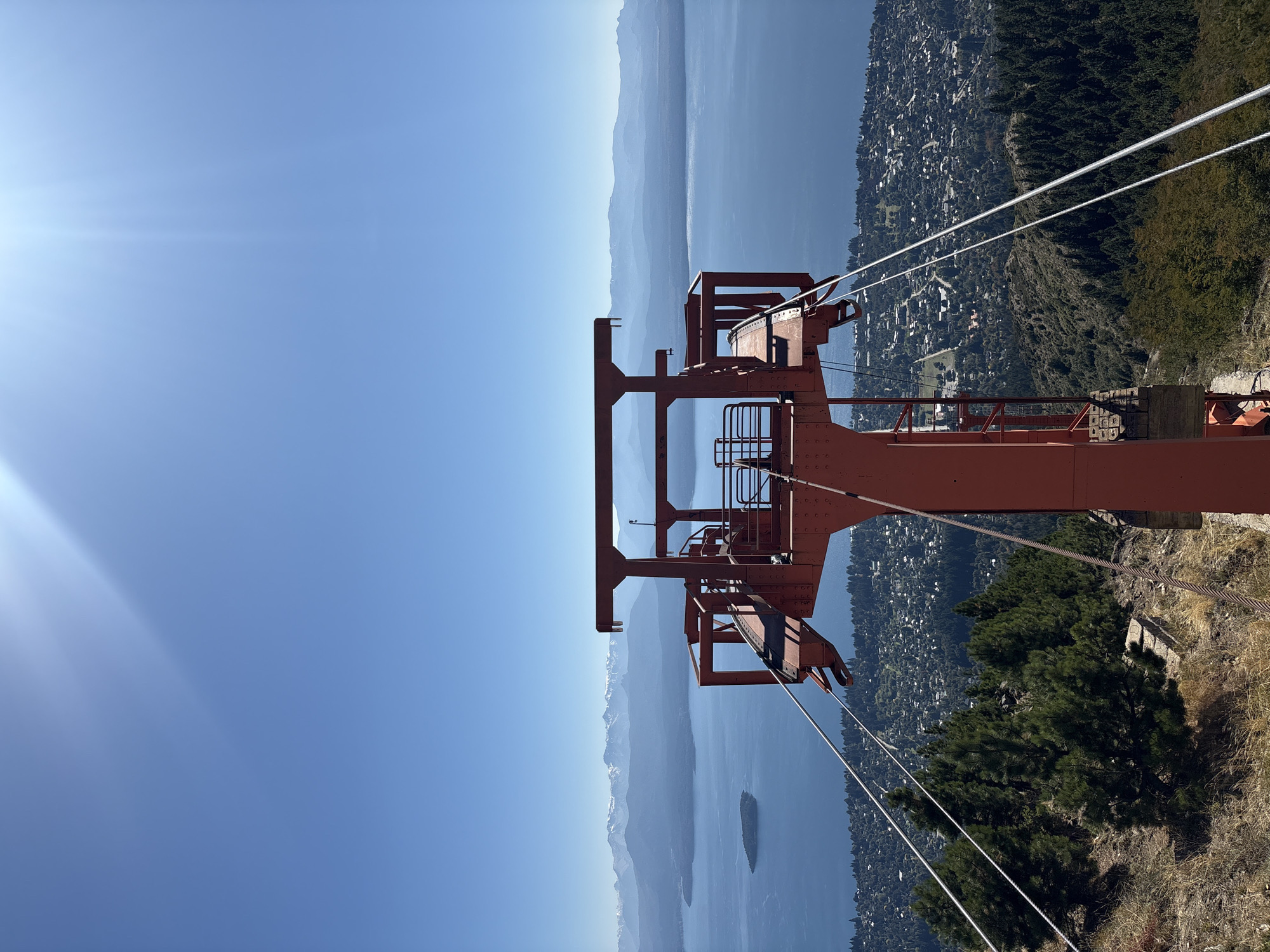 An elevated, static scene captures a panoramic view over San Carlos de Bariloche, Argentina, featuring a prominent, weathered red metal tower in the foreground. This structure, consistent with a cable car or chairlift station, stands centrally, composed of multiple beams, platforms, and railings. Thick, multi-stranded steel cables extend downwards and outwards from the tower, traversing the steep, dry hillside covered with sparse, yellowish grass, exposed earth, and scattered dark green coniferous trees.
Below the immediate foreground, the landscape descends to reveal a dense residential area of San Carlos de Bariloche, characterized by numerous buildings nestled among extensive tree cover. Beyond the city, a large, deep blue body of water, identifiable as Nahuel Huapi Lake, spans the midground. A distinct, oval-shaped, dark green island is visible within the lake towards the left side of the frame.
The background features a series of layered mountain ranges. The closer ranges appear hazy blue, while the furthest peaks exhibit patches of white snow or ice, indicative of the Andean Cordillera. The sky above is a clear, bright cerulean blue, with direct sunlight originating from the upper right quadrant, producing a prominent sunburst or lens flare effect. No individuals or dynamic actions are depicted.