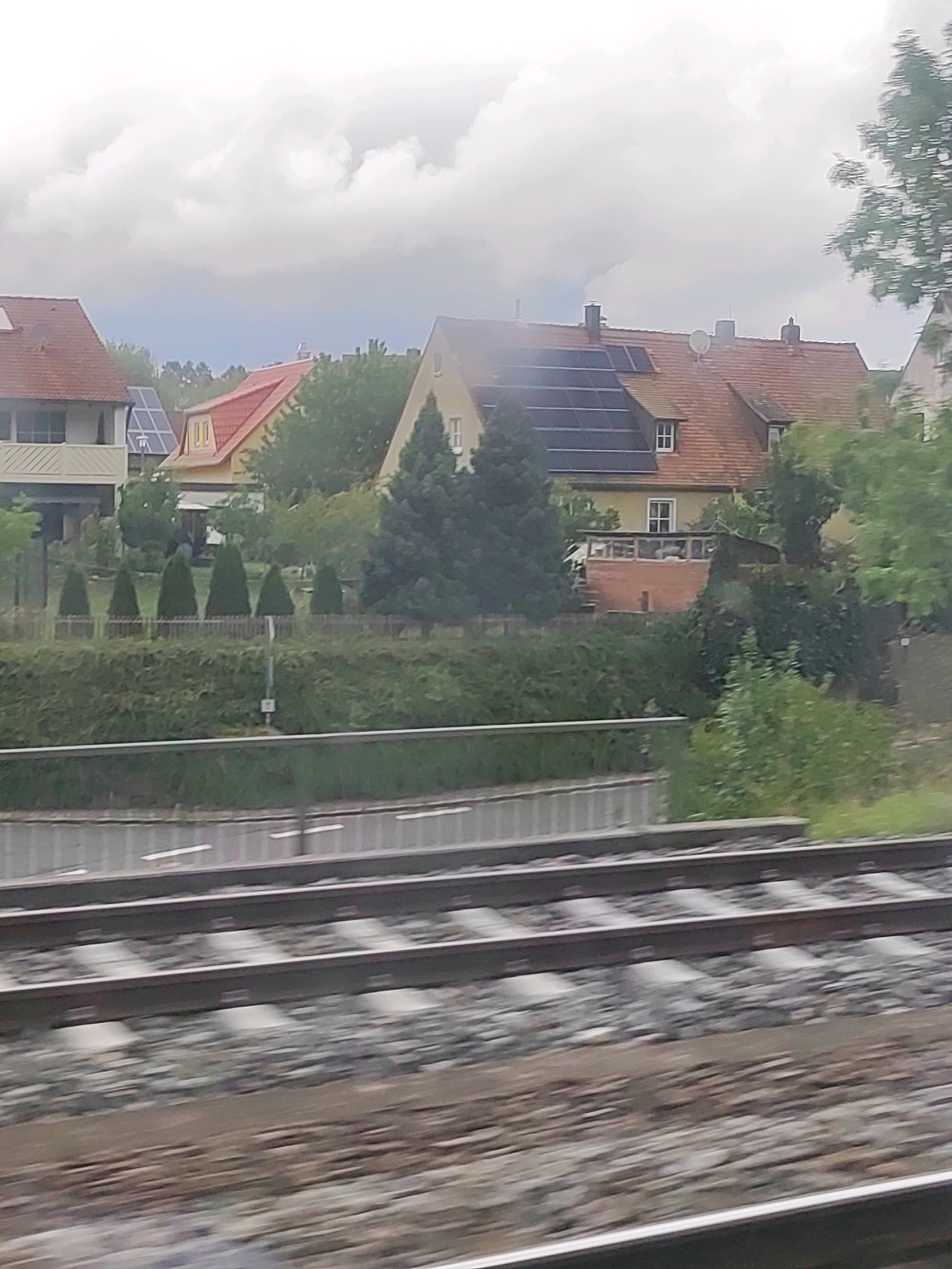 The image shows a blurred outdoor scene with a row of houses and a railway track in the foreground. The houses have red tile roofs, and some have solar panels installed. There are trees and bushes in front of the houses, and a low hedge runs along the property line. A small white sign is visible on a pole behind the hedge. The railway track consists of several parallel sets of rails on ballast. The sky is overcast. The scene is identified as being in Petersaurach, Germany.