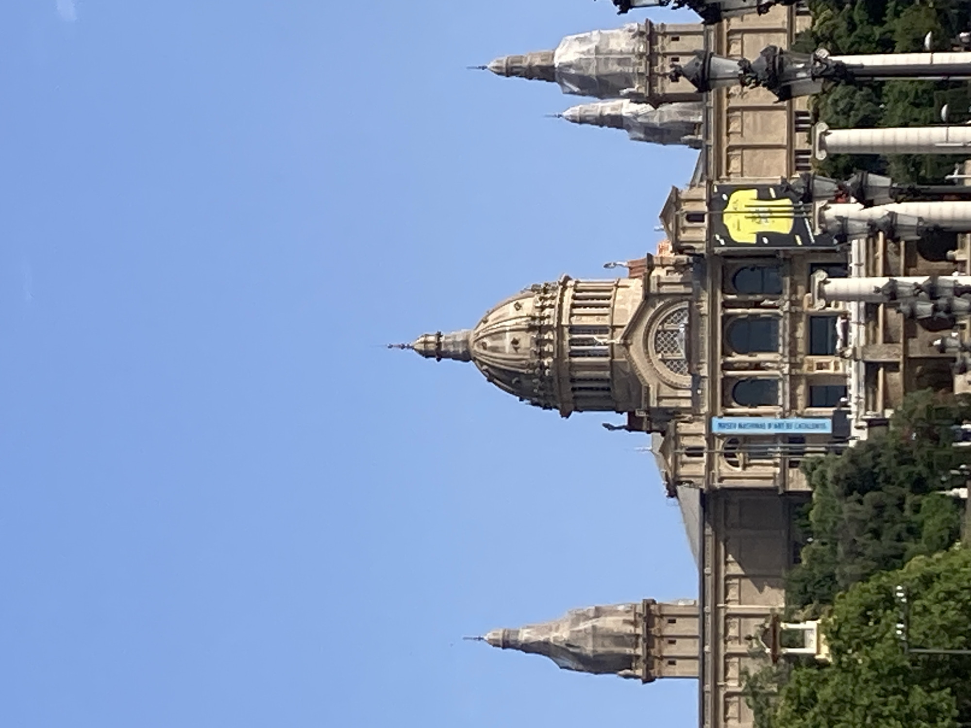 The image shows the Palau Nacional, a grand building in Barcelona, Spain, located on Montjuïc hill. The building features a large central dome, flanked by several towers and spires. The facade is made of stone and is adorned with architectural details. In front of the building, there are trees and a clear blue sky. Two banners are visible hanging from the building, one blue and one yellow with a graphic design. A set of decorative street lamps are in the foreground.