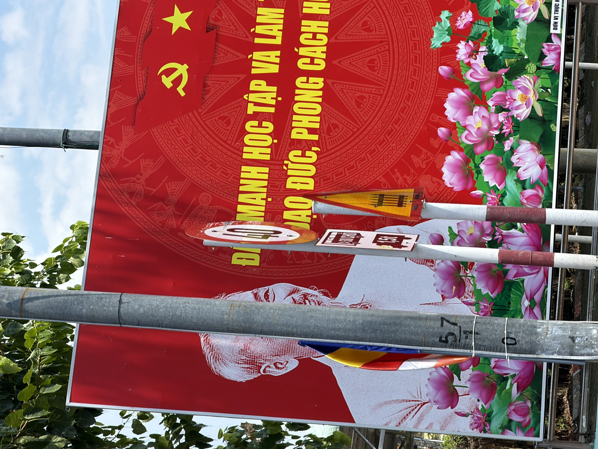 A large, red billboard in Tay Nha Trang Ward, Vietnam, features a portrait of a man with white hair and a beard, rendered in white. The billboard is adorned with traditional Vietnamese patterns and a yellow hammer and sickle symbol, alongside a yellow star. Text in yellow Vietnamese characters reads "MANH HOC TAP VA LAM" and "DAO DUC, PHONG CACH HO". The lower portion of the billboard displays an arrangement of pink lotus flowers and green leaves. A gray metal pole stands in front of the billboard, obscuring parts of the image. A small Vietnamese flag with red and yellow stripes is attached to the pole. Several traffic signs are visible on the right side of the billboard, including a circular sign with "40" and a rectangular sign with "1.51". A metal fence runs along the bottom of the image. Trees and a cloudy sky are visible in the background.