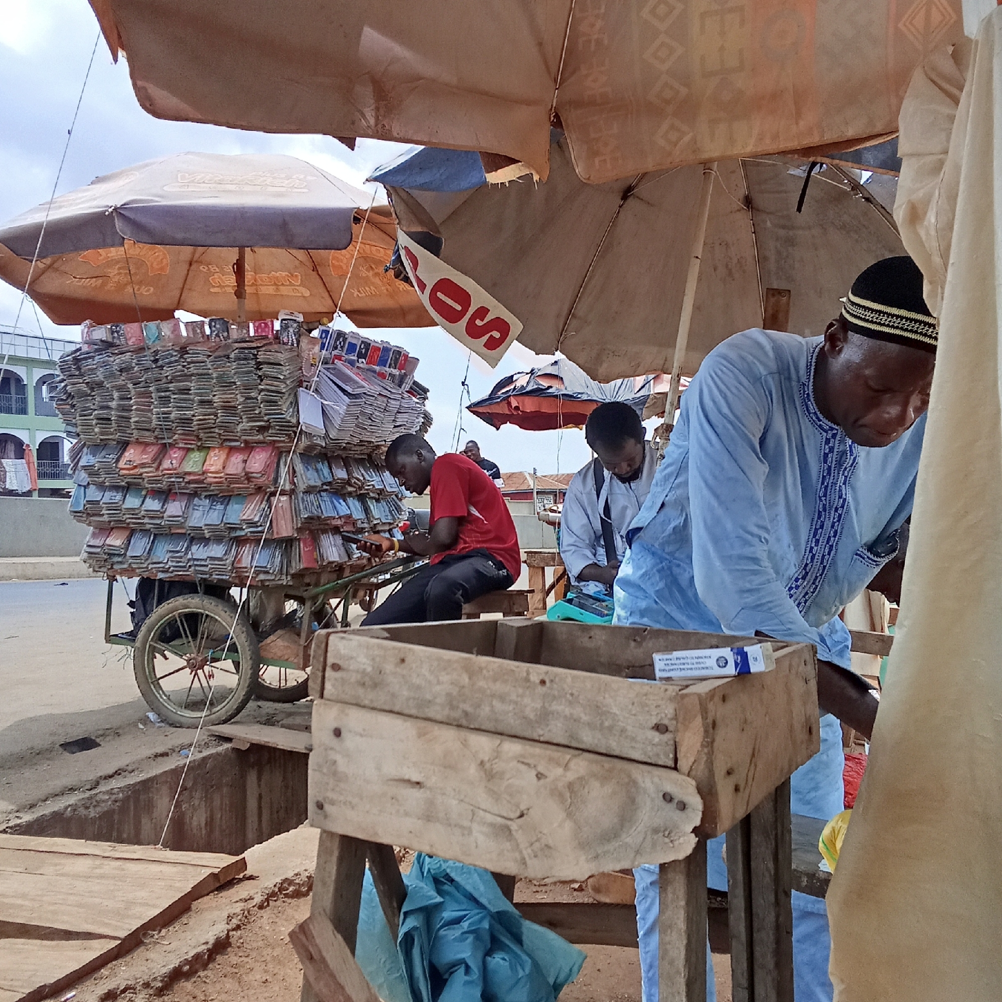 This image depicts a street scene in Akwanga, Nigeria, showcasing a mobile vendor of what appears to be mobile phone accessories and covers. The vendor's wares are stacked high on a wheeled cart beneath several large sun umbrellas. Two men sit on the cart's frame, and a third man wearing a blue robe and a traditional cap is positioned in front of a wooden crate, likely handling transactions. The scene is somewhat cluttered, with umbrellas providing shade, and the background shows buildings and the urban environment. The image is taken at ground level and captures a slice of everyday commercial activity in Akwanga.